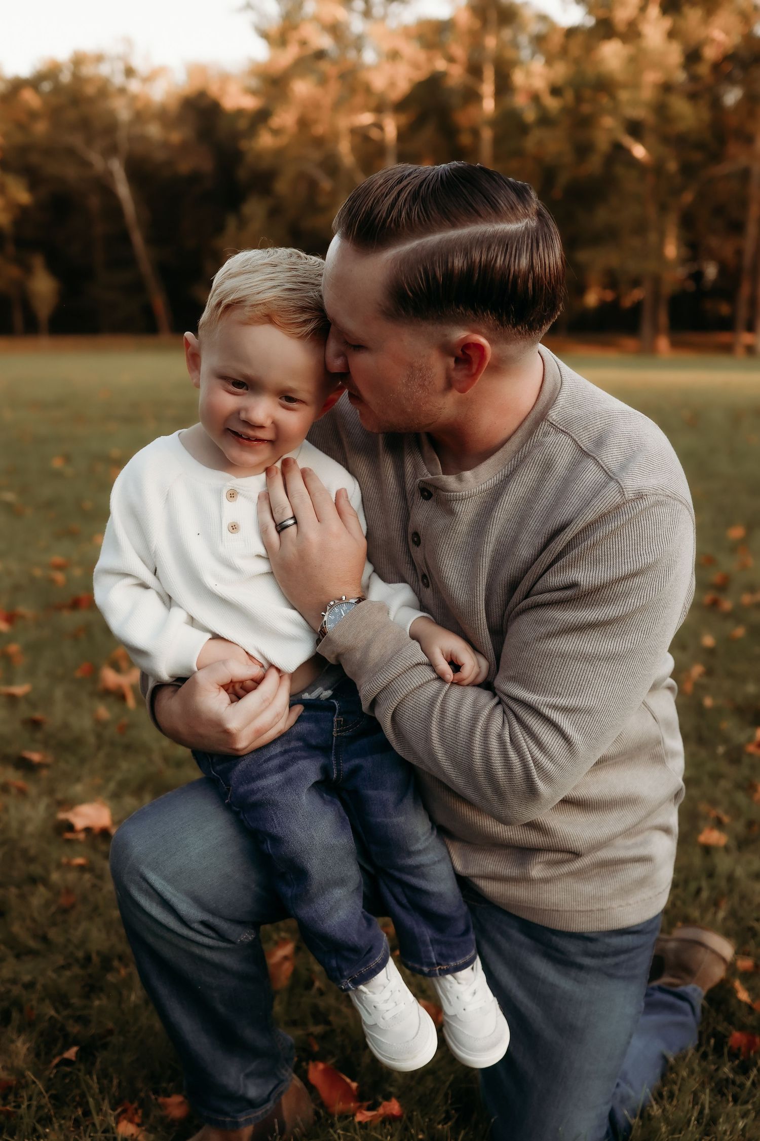 Affectionate father kissing smiling toddler son on cheek in fall outdoor setting.