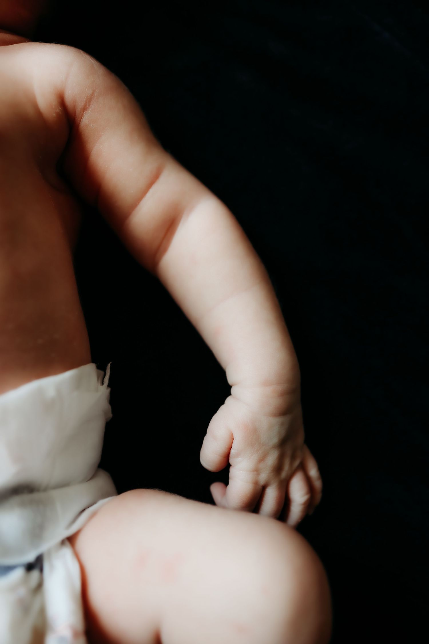 Detailed close-up of a newborn baby's arm and hand resting on a diaper against dark background.