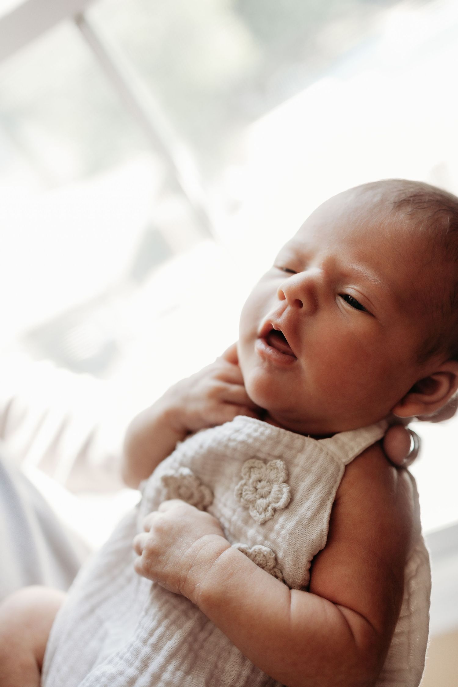 Peaceful newborn baby in white floral muslin romper being held upright in soft natural light.