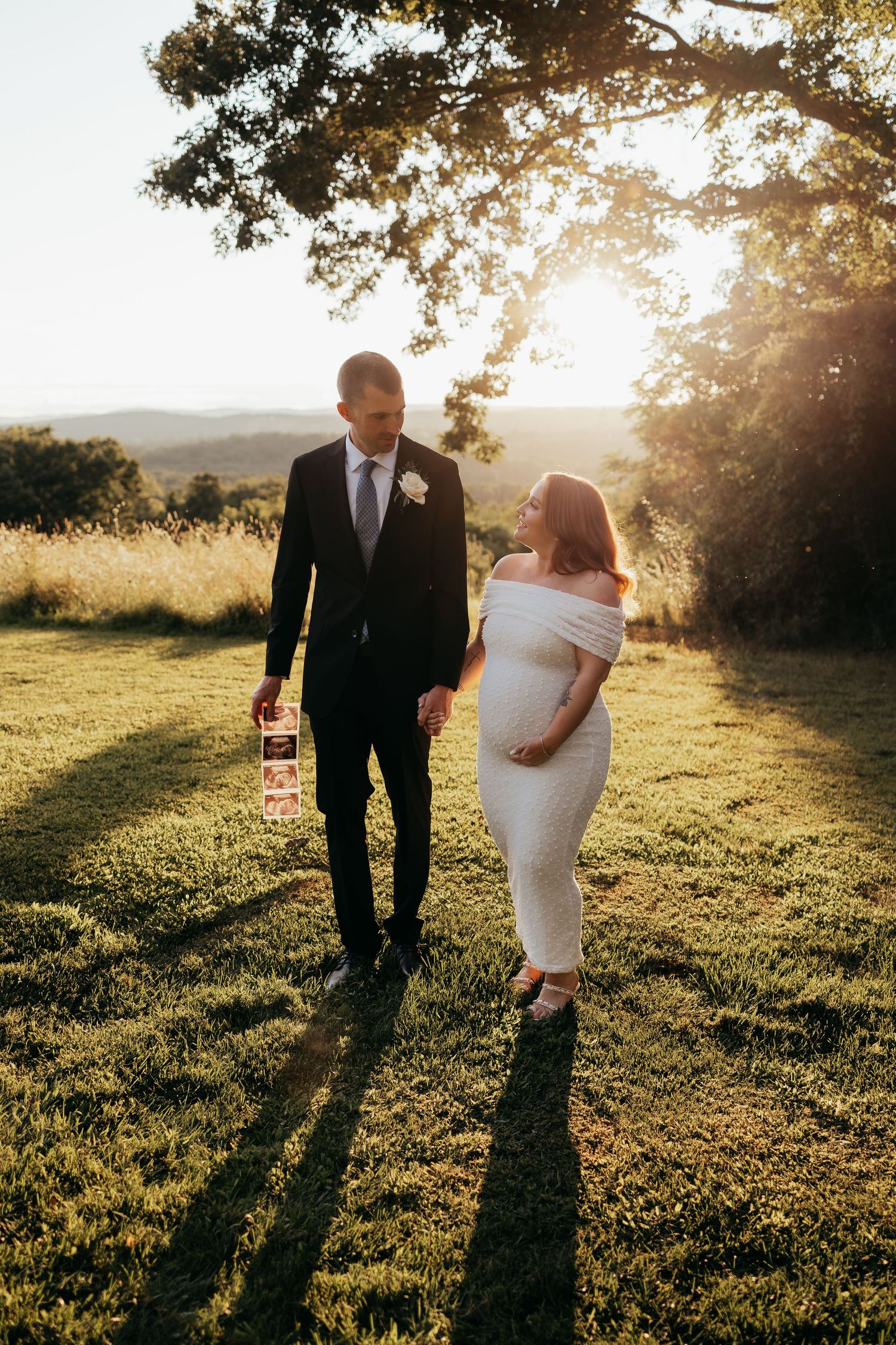 Pregnant bride in white off-shoulder dress holds hands with groom in black suit at golden hour outdoors.