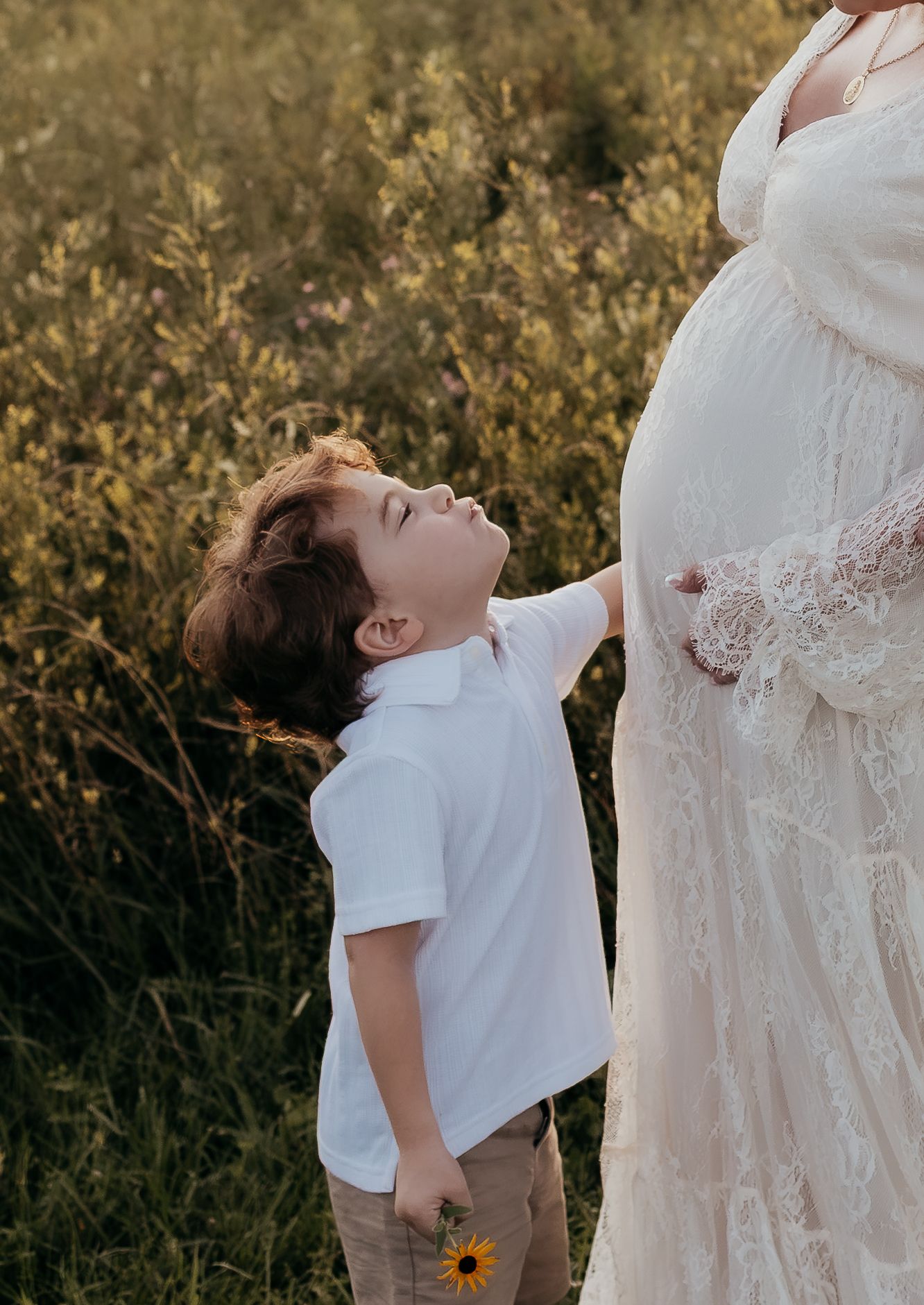 A young boy in a white polo touches a pregnant woman's belly in a sunlit field, holding a yellow sunflower.