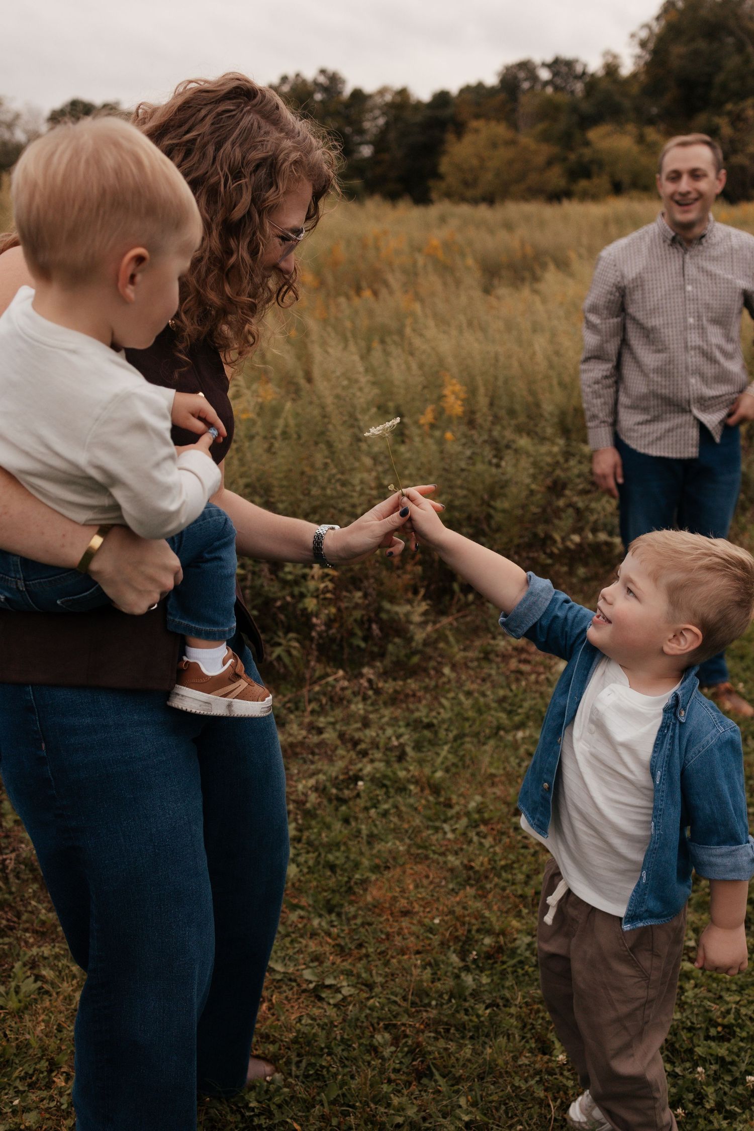 Family of four enjoying an autumn day in a field, mother holding baby while toddler reaches out to touch hands.