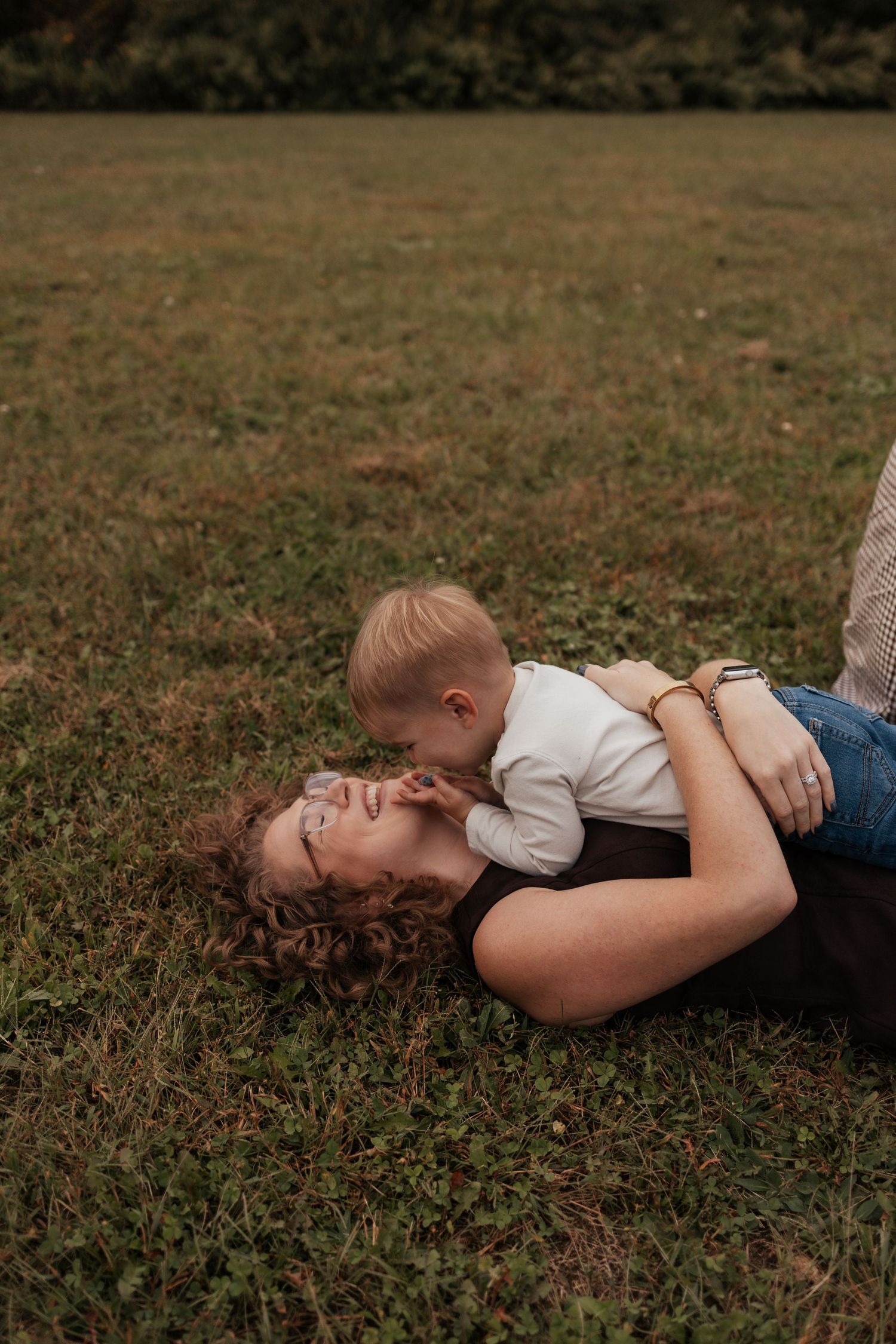 A mother lying on green grass while her toddler leans in for a kiss, sharing a tender outdoor moment.