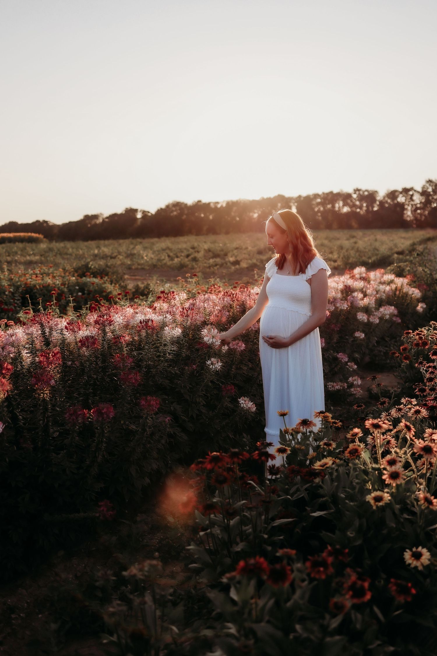 Pregnant woman in white dress standing in a vibrant flower field at golden hour, cradling her baby bump.