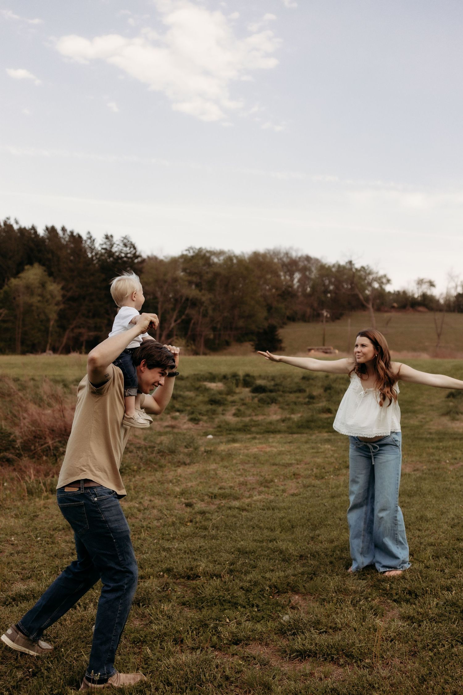 Family playing in a green field, father carrying toddler on shoulders while mother reaches out joyfully.