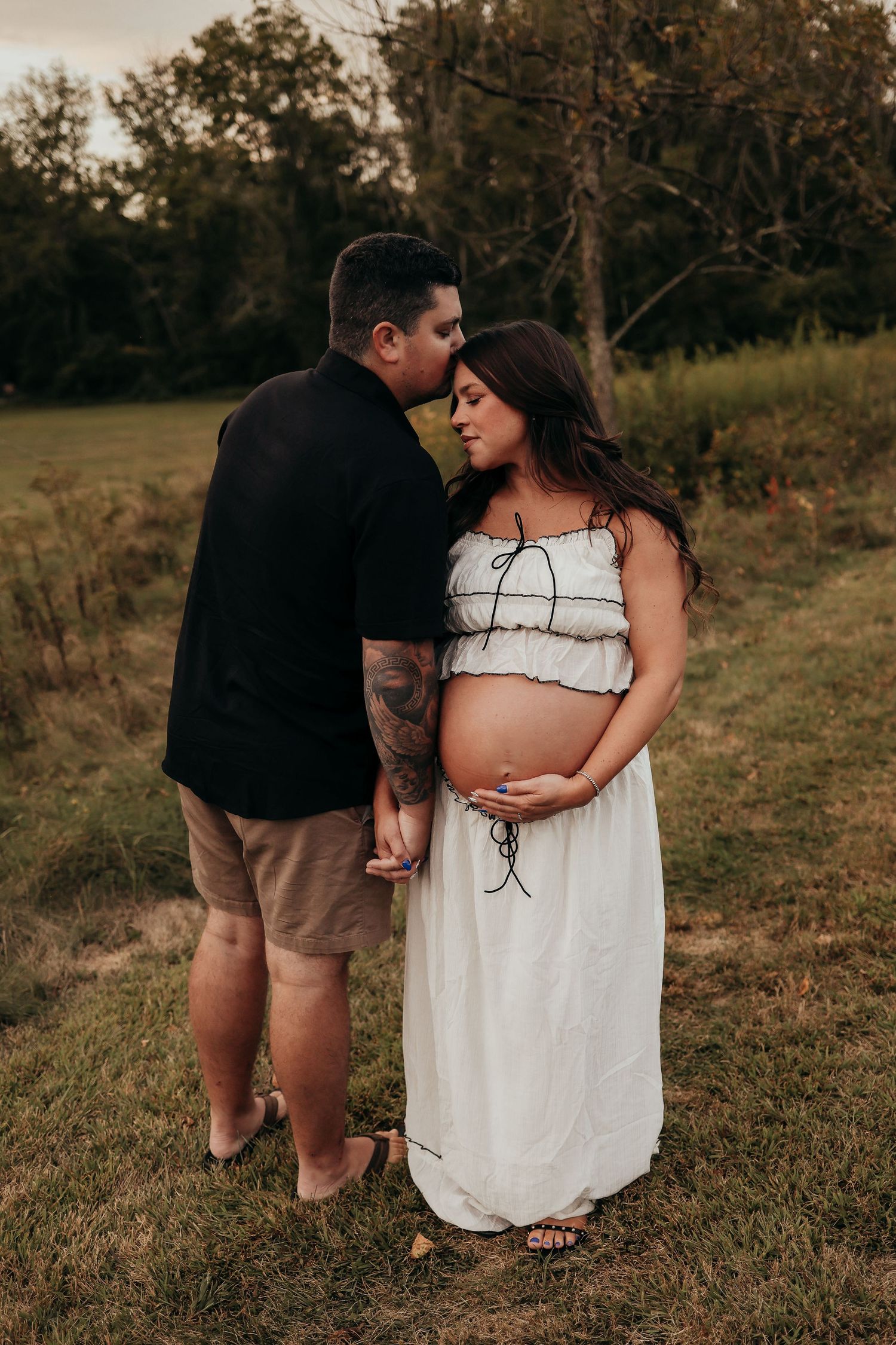 Tattooed man kissing pregnant woman's temple while holding hands in a grassy field at golden hour.
