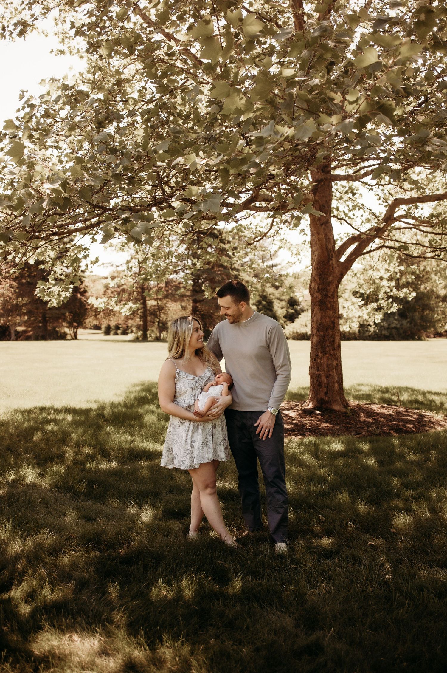 Young couple standing under large tree in sunny park lovingly gazing at their newborn baby.