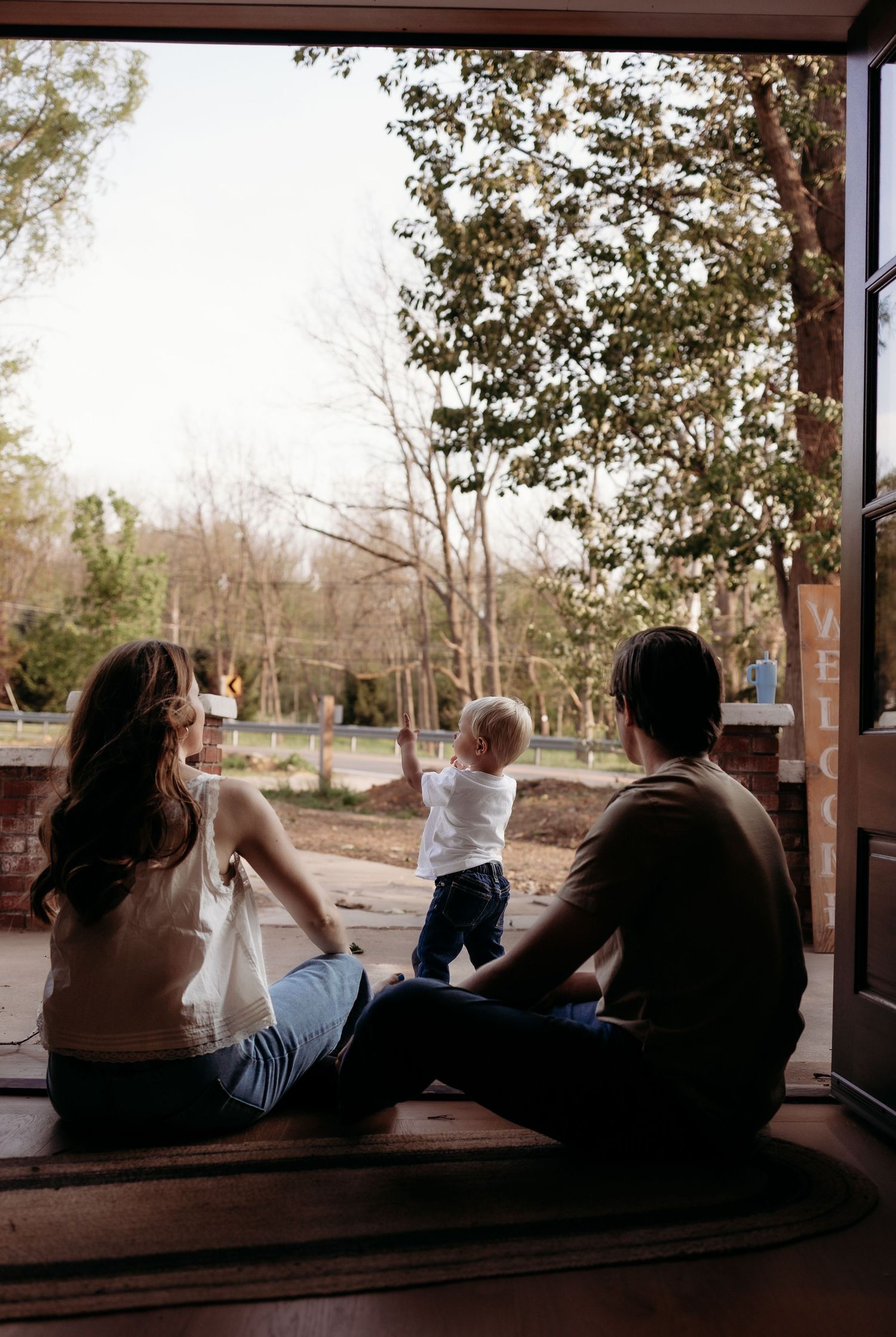 A young family sits in an open doorway watching their toddler play outside surrounded by trees.