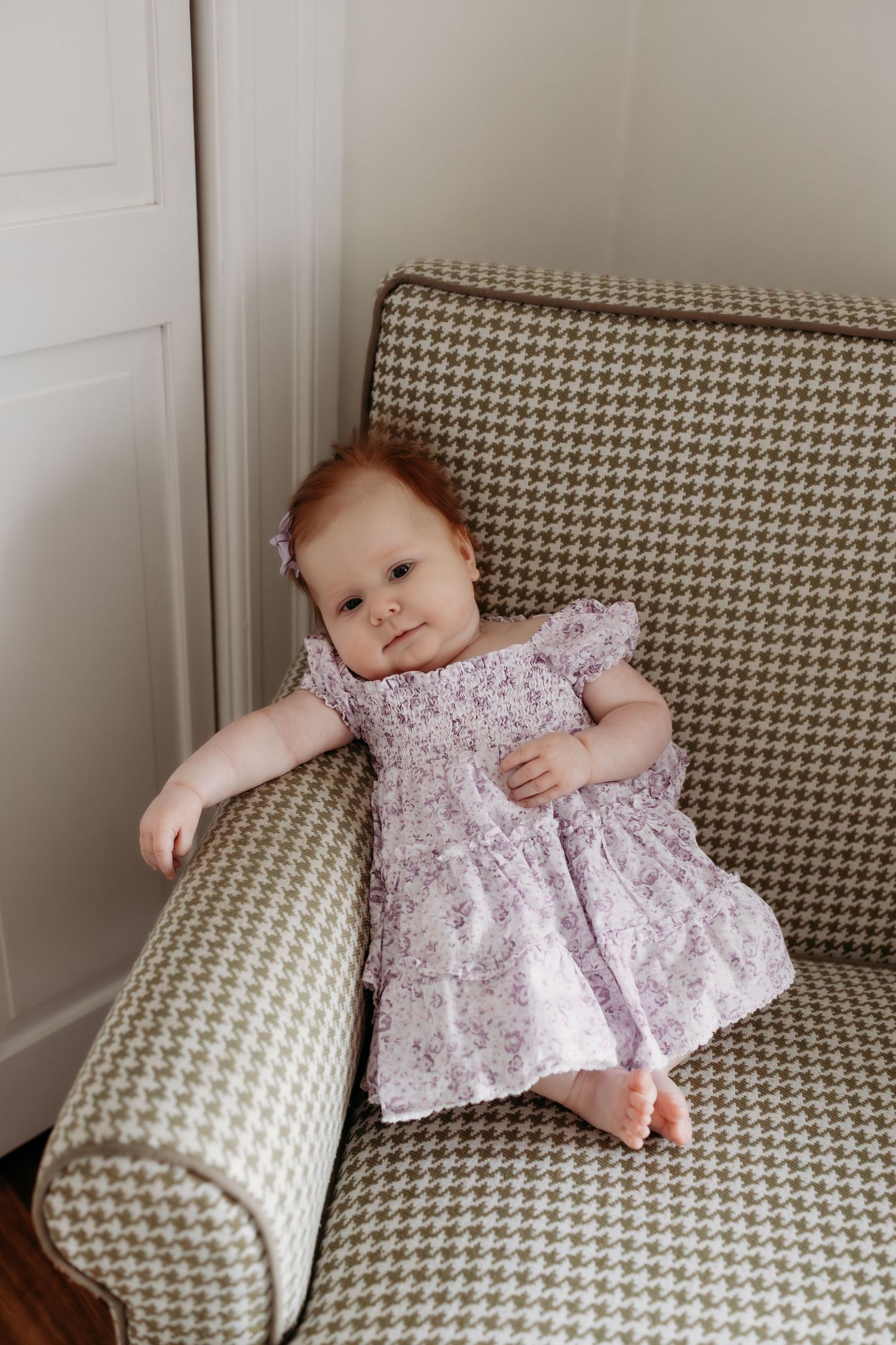 Baby girl in floral purple dress sitting on houndstooth armchair, looking at camera.