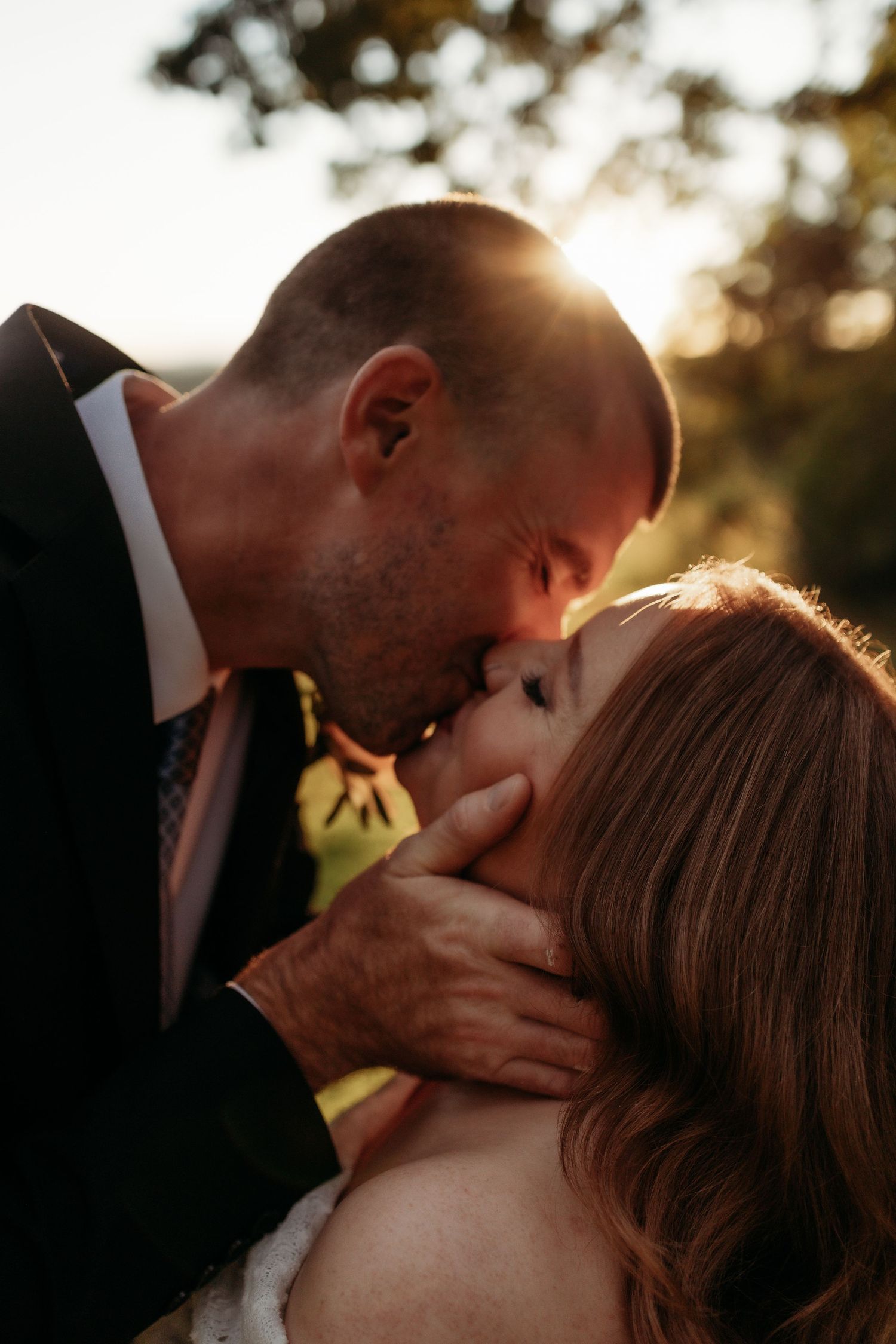 Groom tenderly kissing bride at golden hour, hand gently cradling her face outdoors.