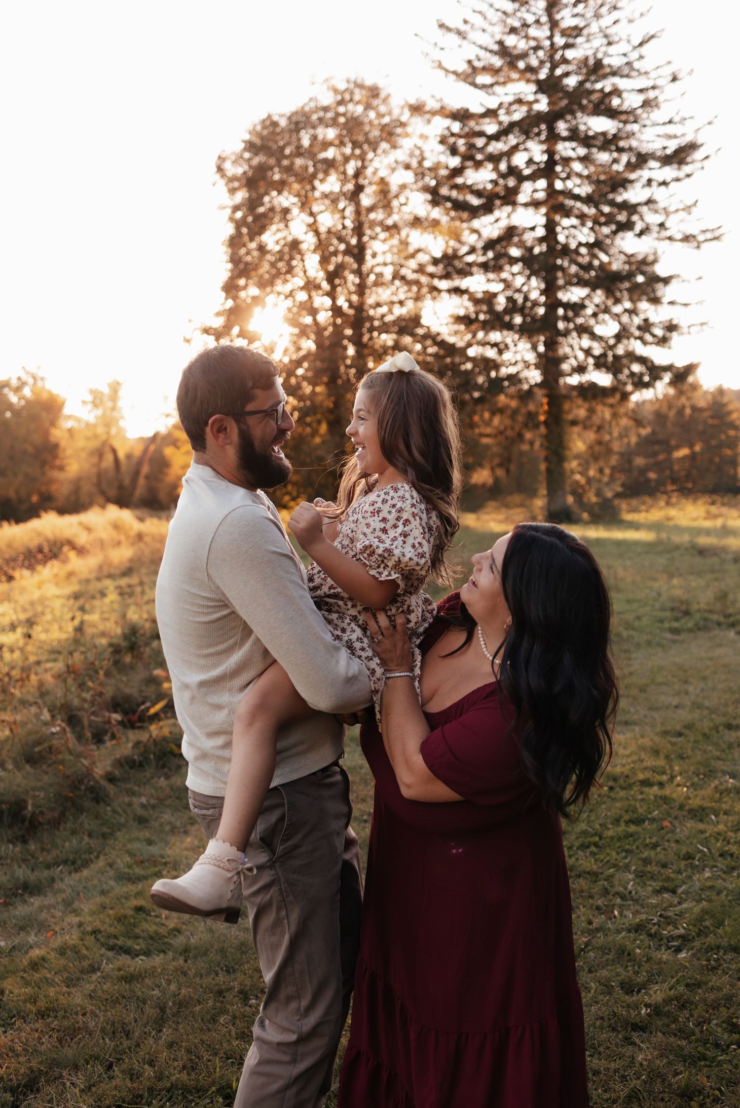 A smiling family of three shares a joyful moment outdoors at golden hour, with a father lifting his young daughter beside her mother.