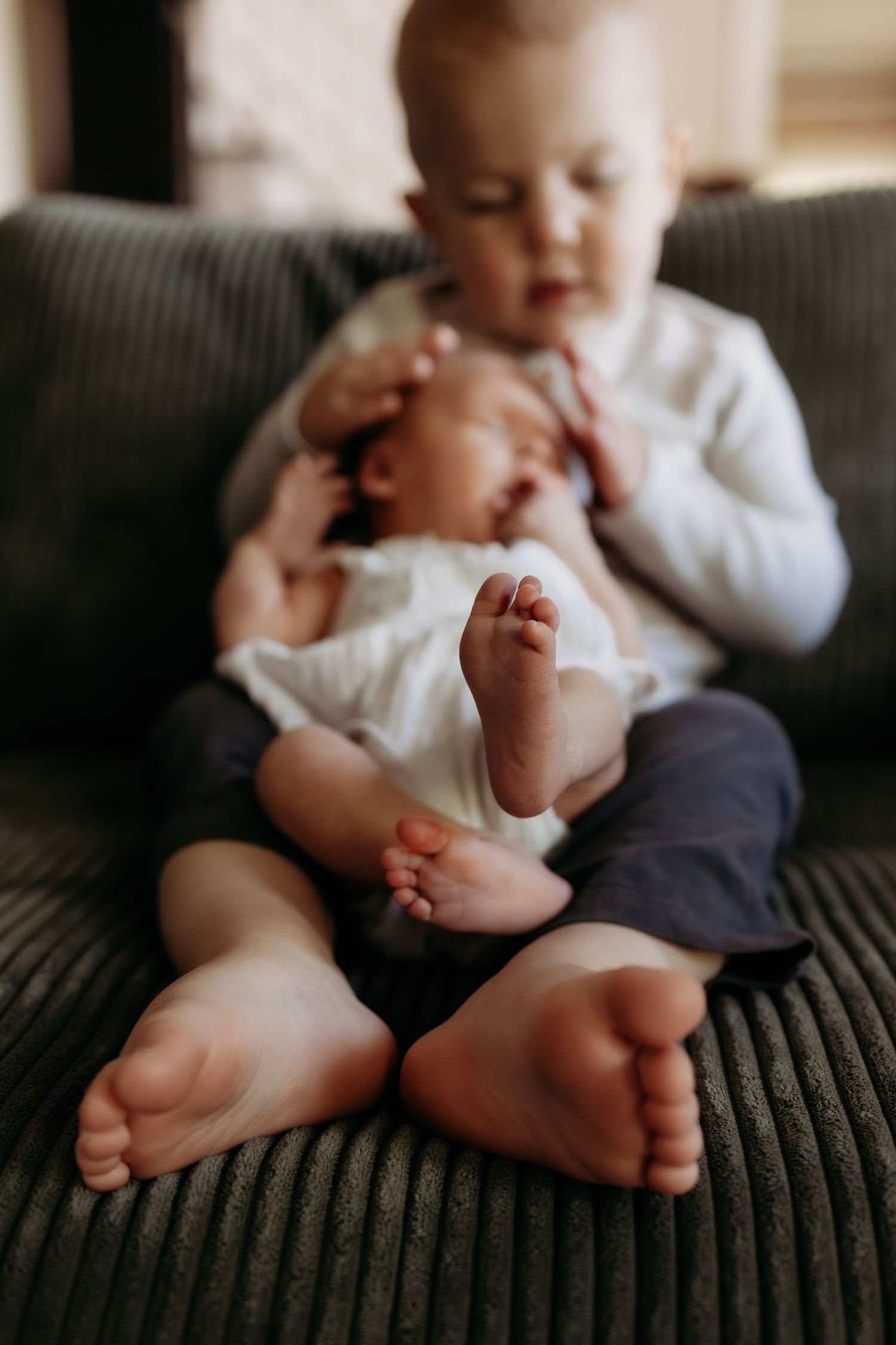 Toddler sibling gently holding sleeping newborn baby on gray sofa, tiny feet in focus.