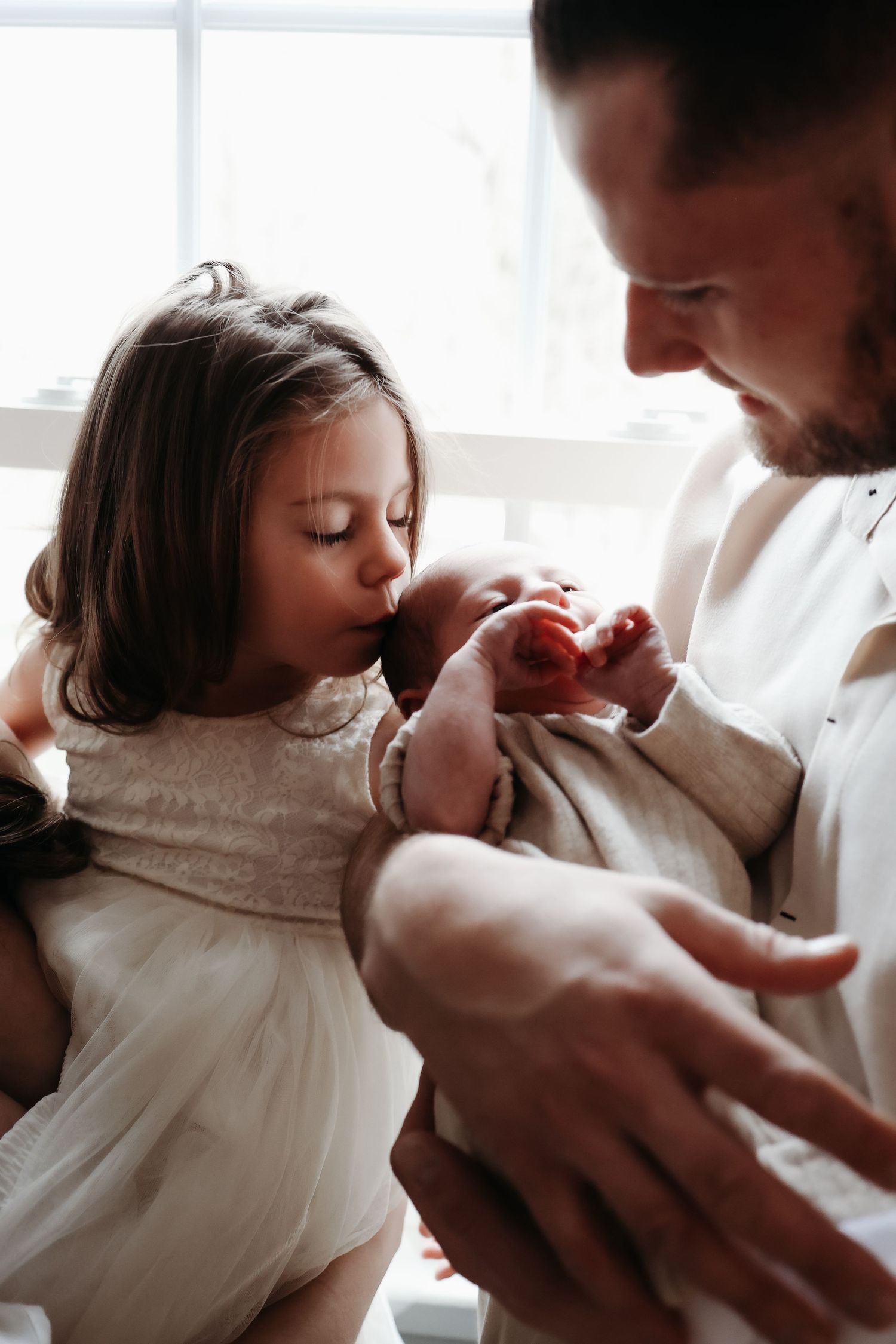 A young girl gently kisses a newborns hand while father holds the baby near a bright window.