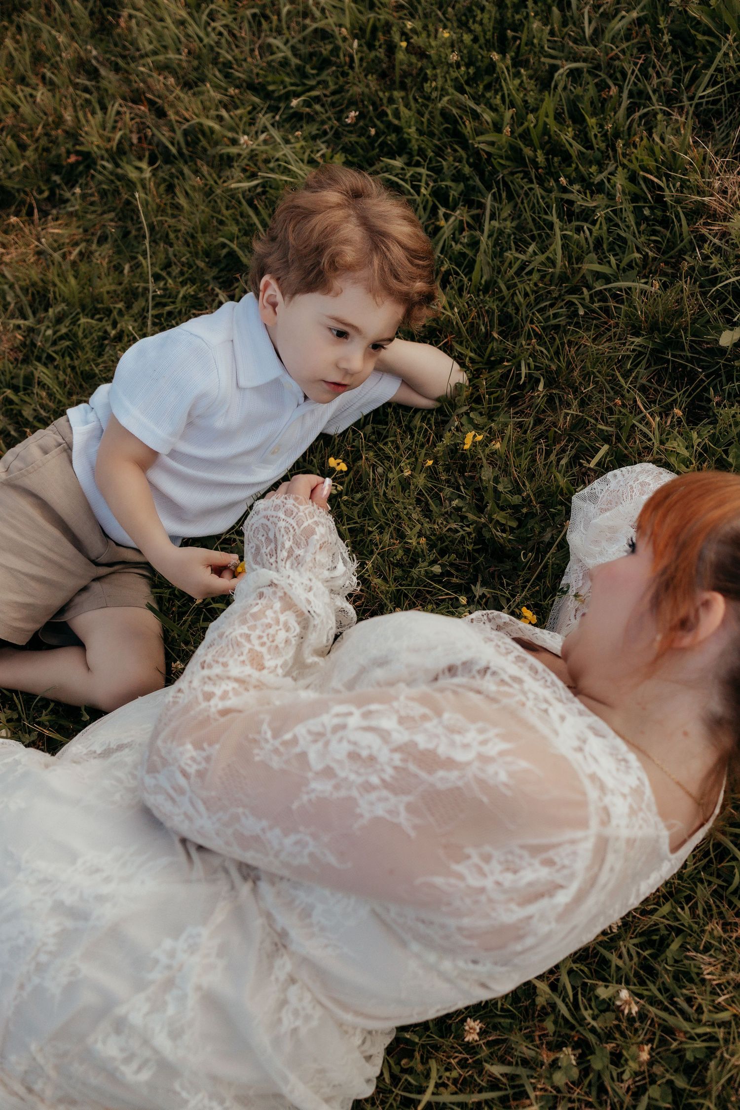 A mother in a white lace dress lies on grass with her young redheaded toddler son during an outdoor maternity photoshoot.