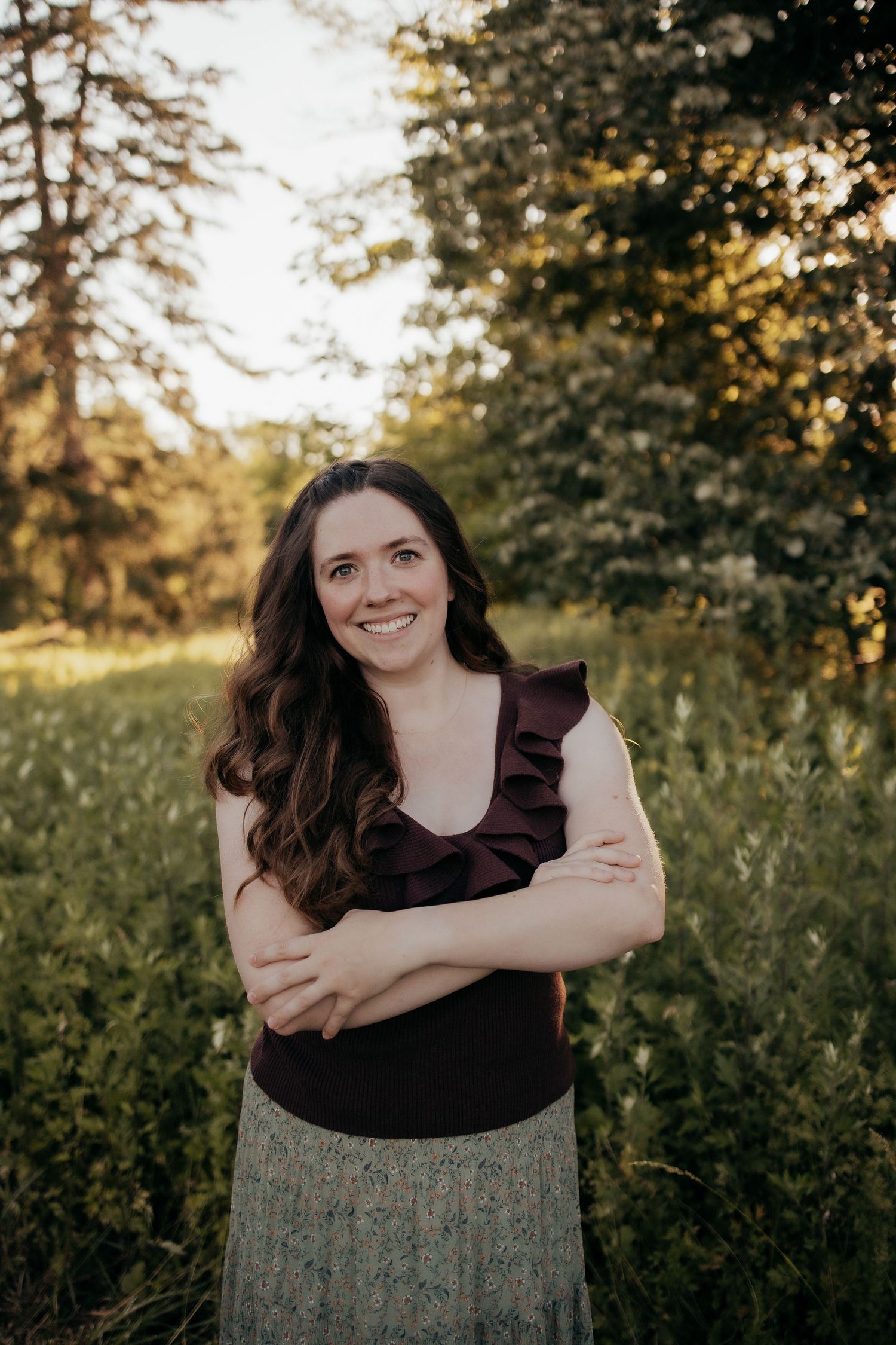 Cheerful woman in dark ruffled sleeveless top smiles with arms crossed in a sun-drenched green meadow.