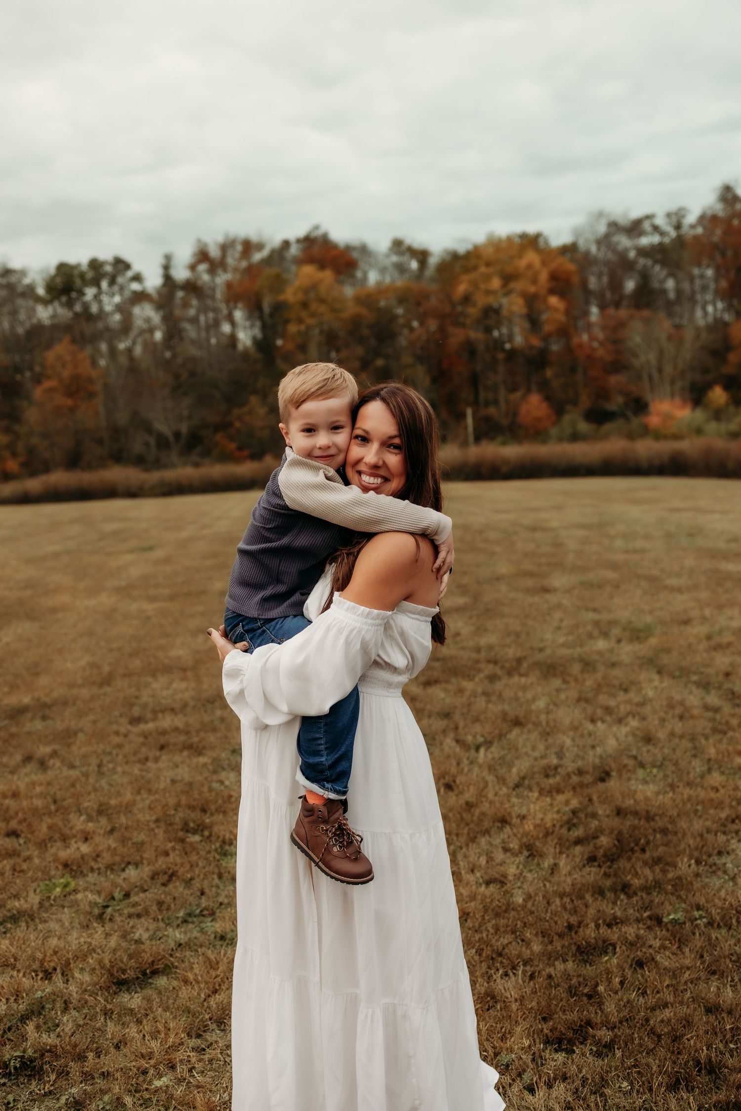 Woman in white dress holding toddler boy in autumn field.