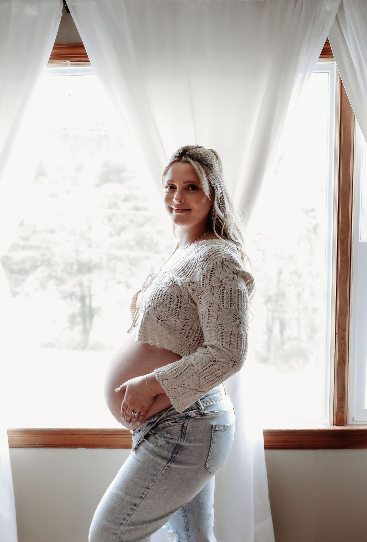 Pregnant woman in cream knit crop top and jeans posing by a bright window.