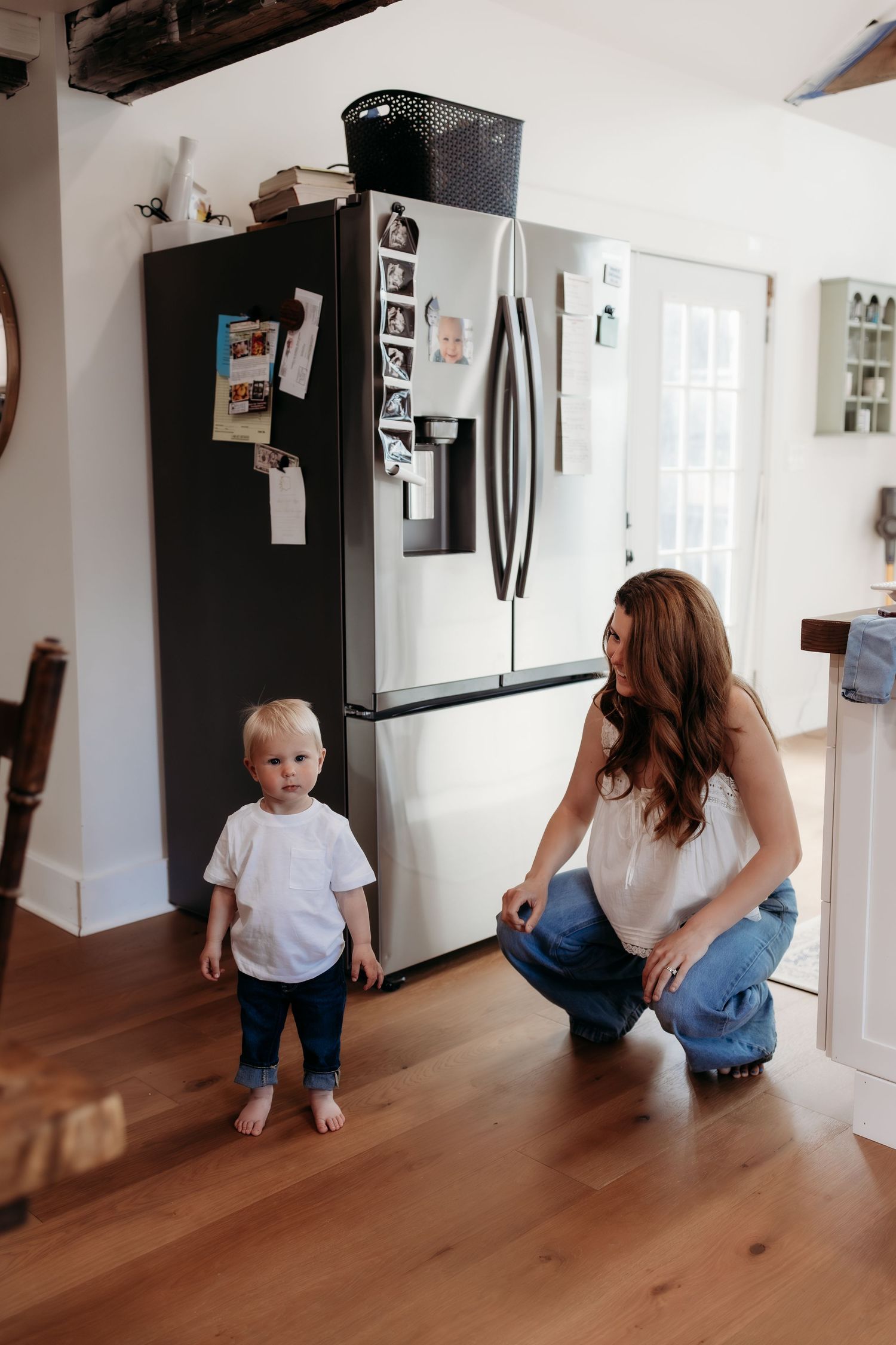 Mother kneels smiling at barefoot toddler standing in modern kitchen.
