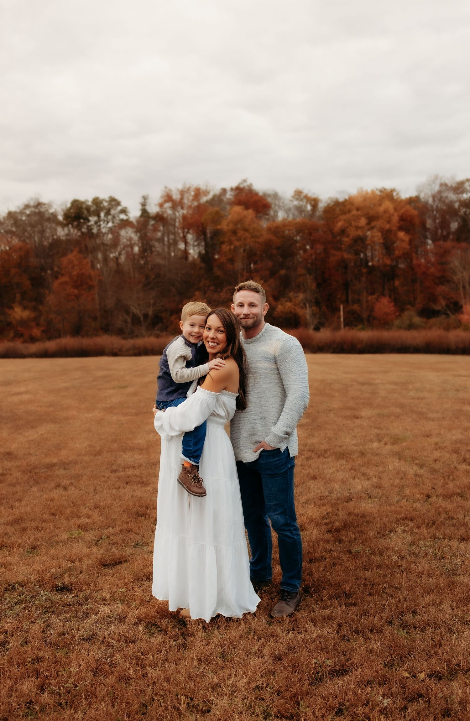 Family portrait in autumn field with expecting mother in white dress and young child against backdrop of orange fall foliage.