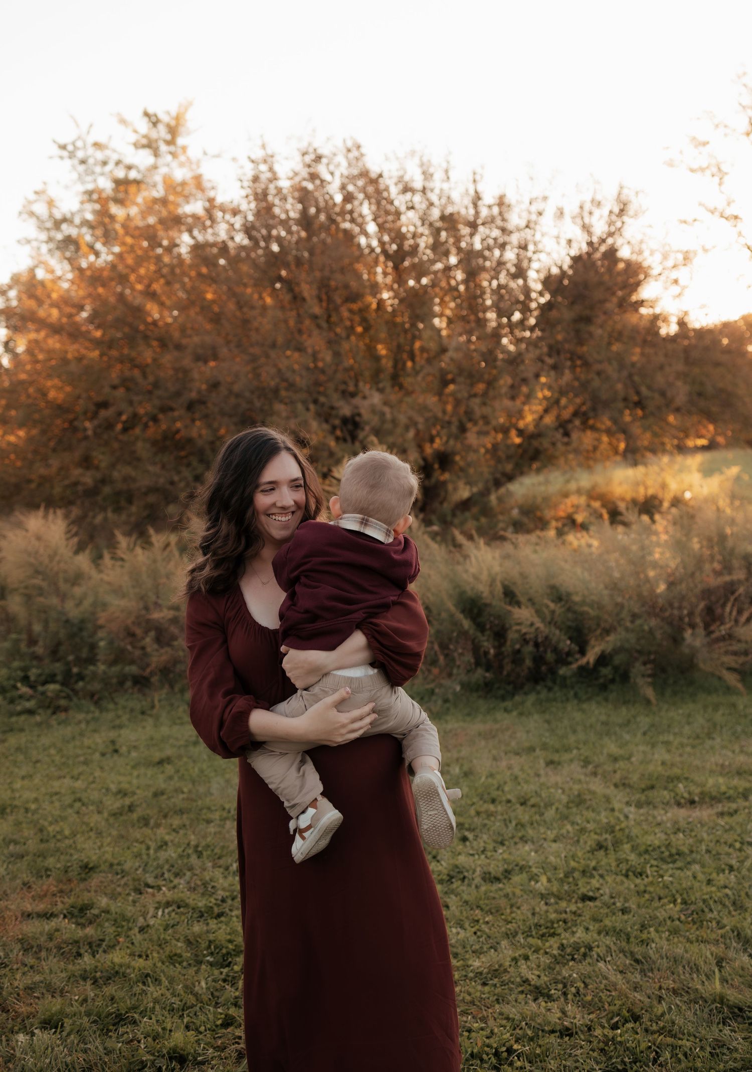 A smiling woman in a burgundy dress holds a toddler in her arms in a grassy field at golden hour in autumn.