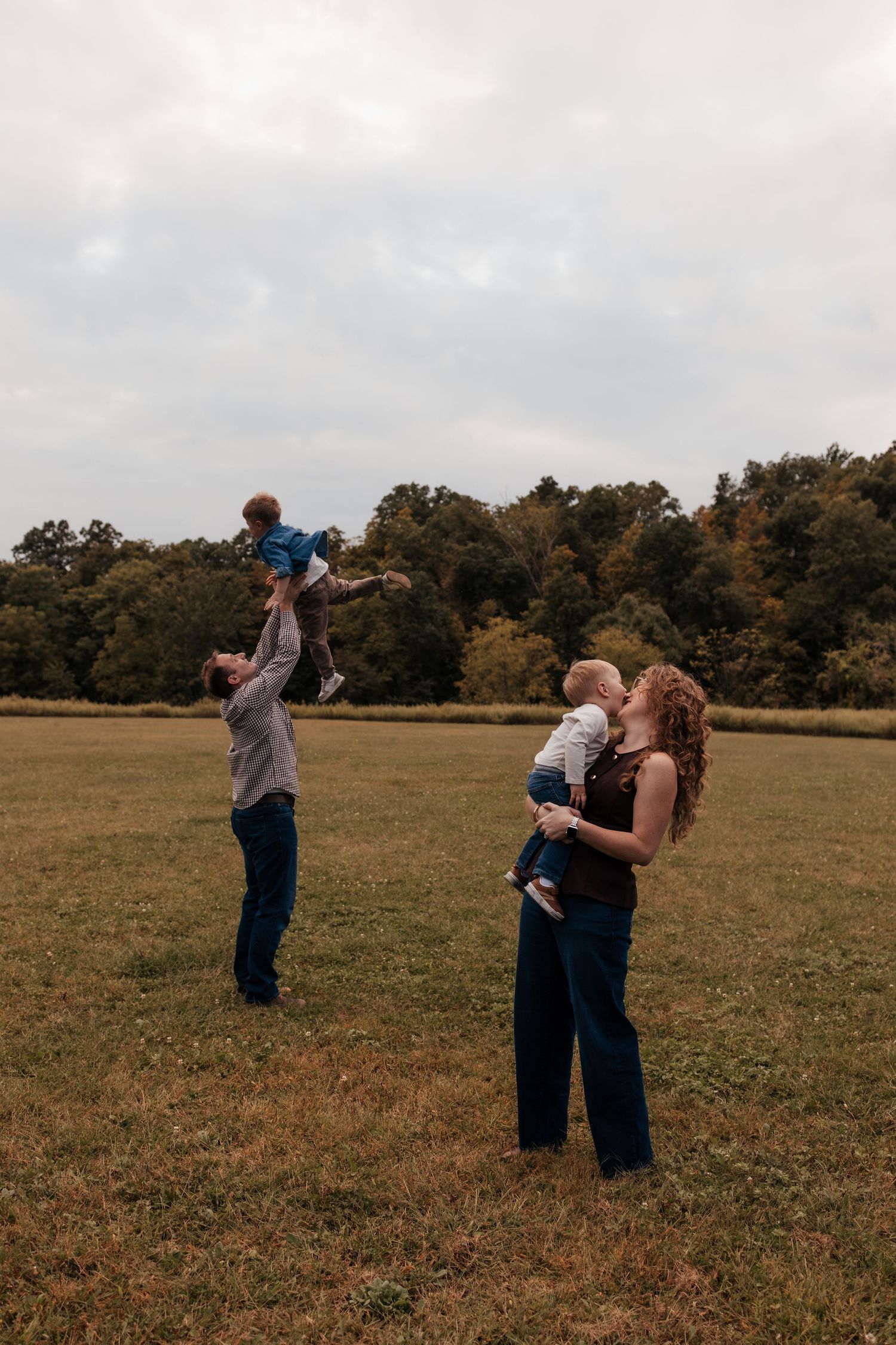 Family playing outdoors, father tossing child in air while mother kisses baby.