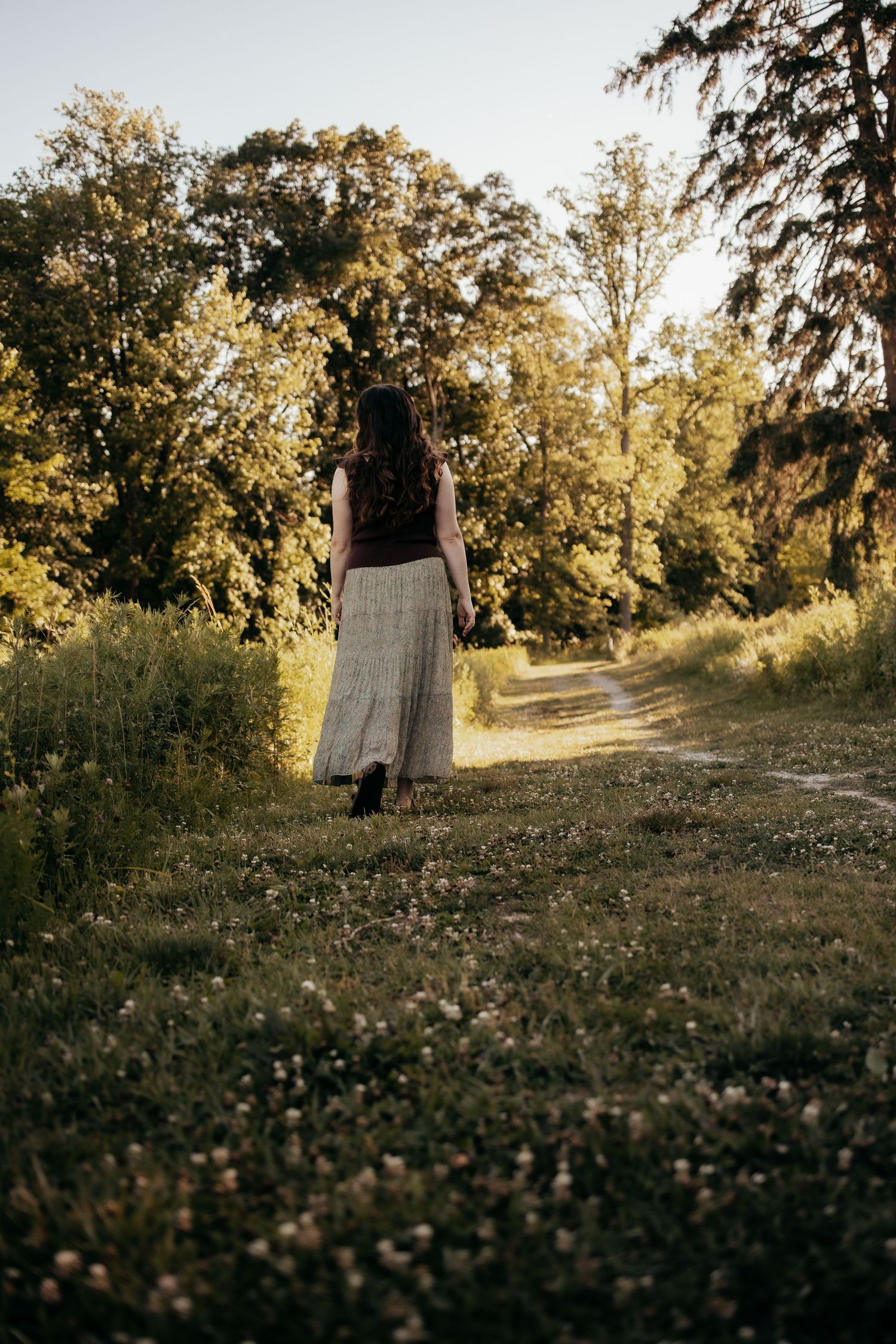 Woman in maxi skirt walking along sunlit tree-lined path in lush green landscape.