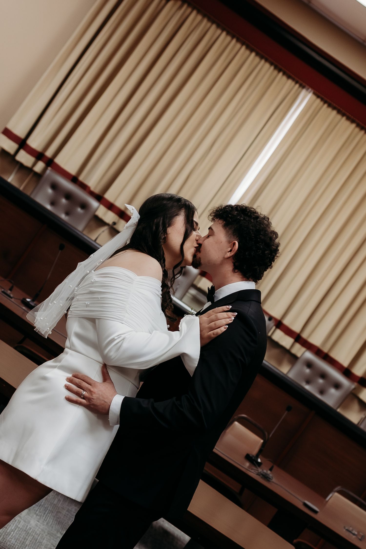 Bride and groom sharing first kiss in an indoor ceremony room, bride in white off-shoulder mini dress.