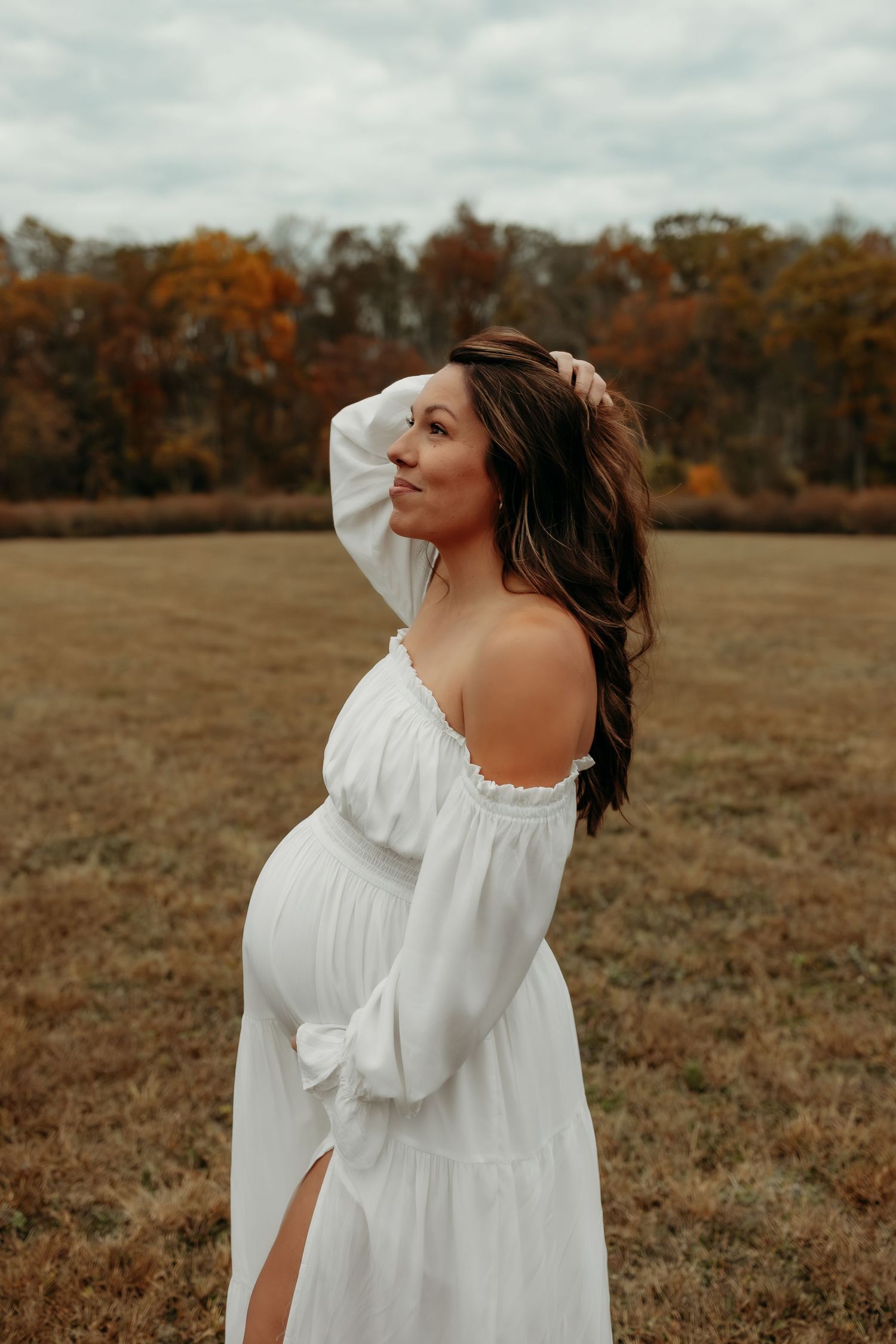Pregnant woman in white off-shoulder dress posing in autumn field.