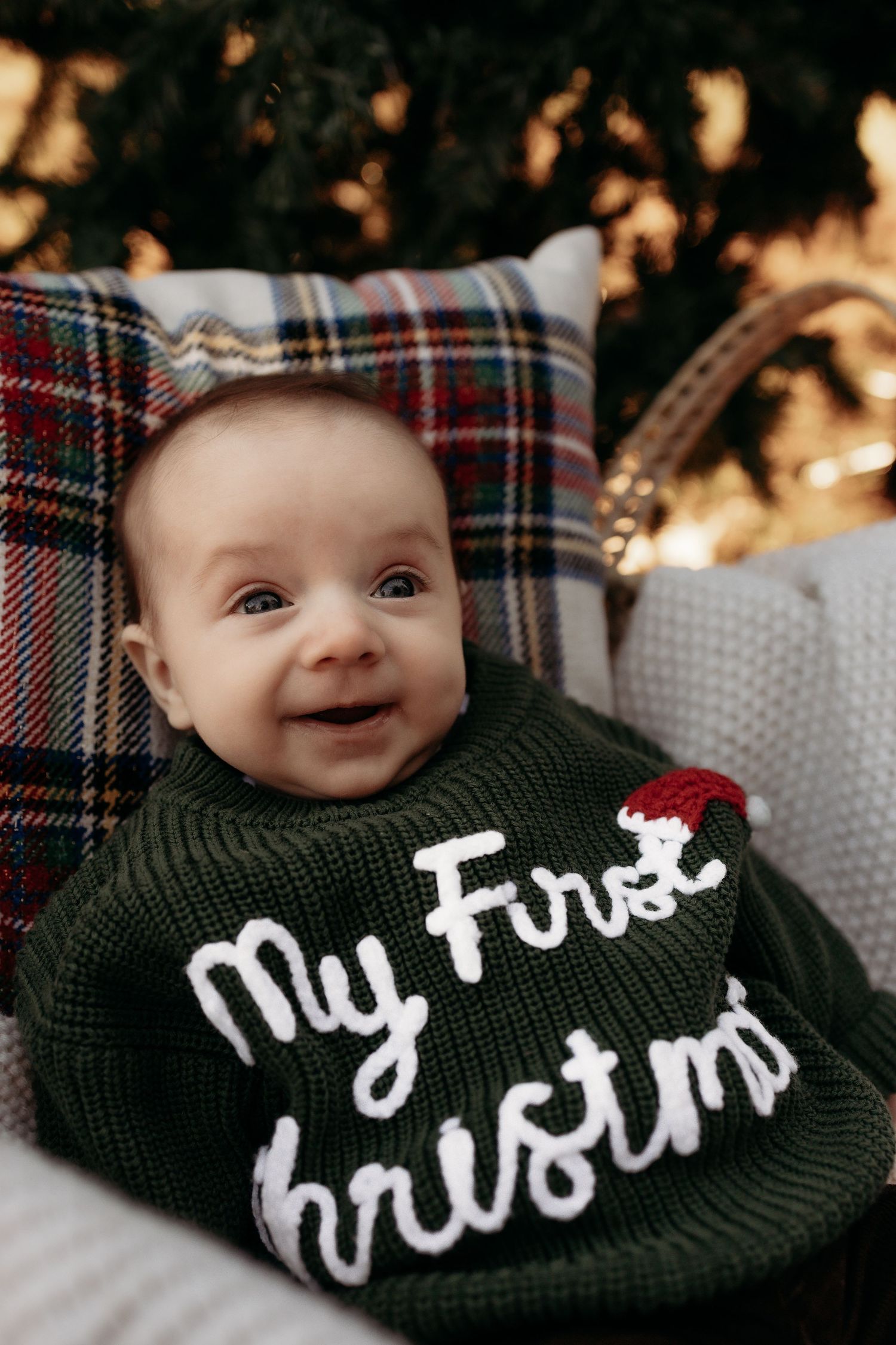 Smiling baby in a green My First Christmas sweater, nestled in a basket with plaid blanket and holiday tree backdrop.