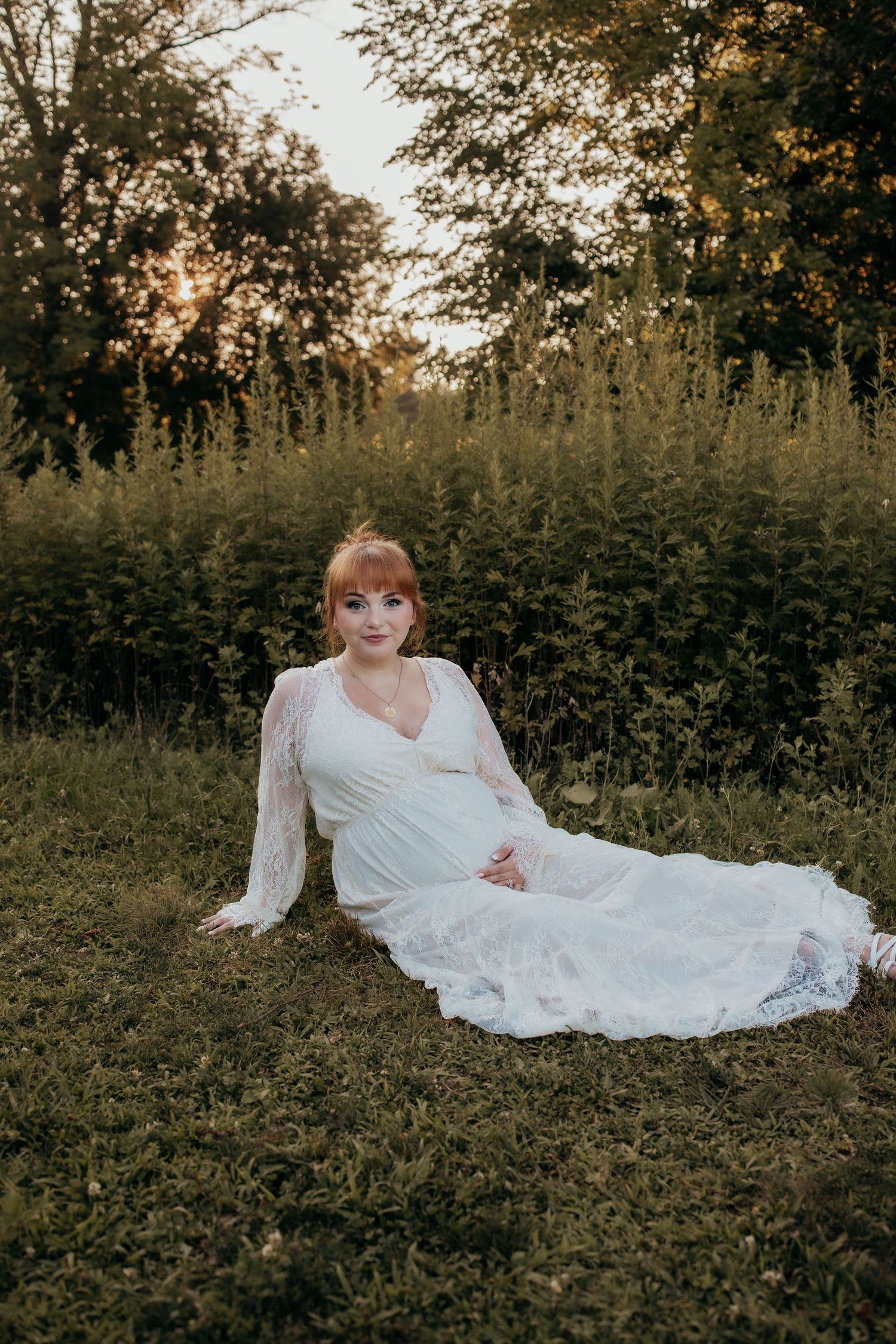A woman in a white lace gown sits gracefully on green grass at sunset with lush trees in the background.