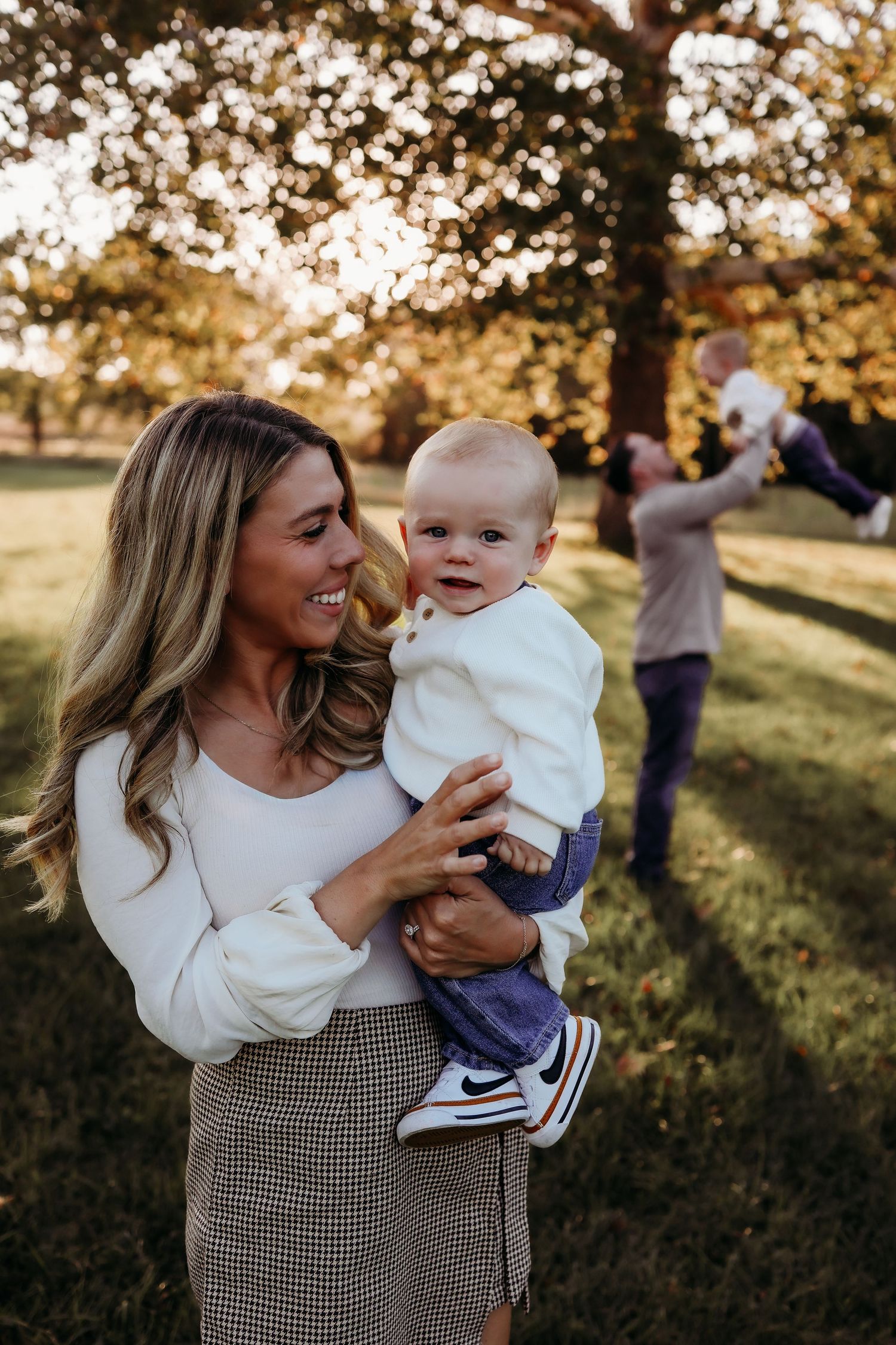 Smiling mother holding baby boy outdoors while father plays with toddler in background.