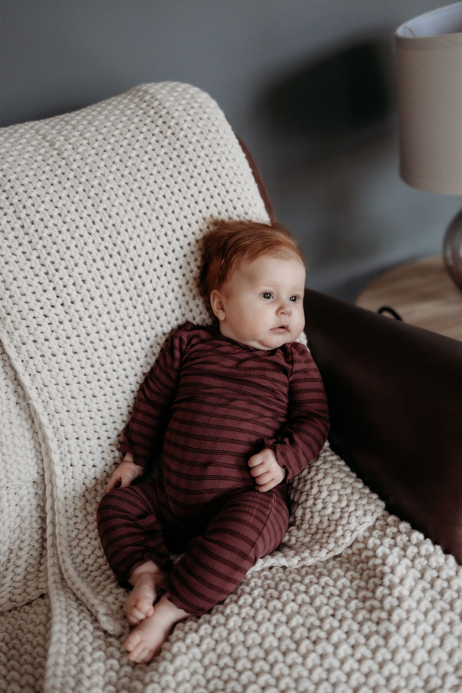 Adorable red-haired baby in burgundy striped pajamas resting on a cream knit blanket on a cozy armchair.