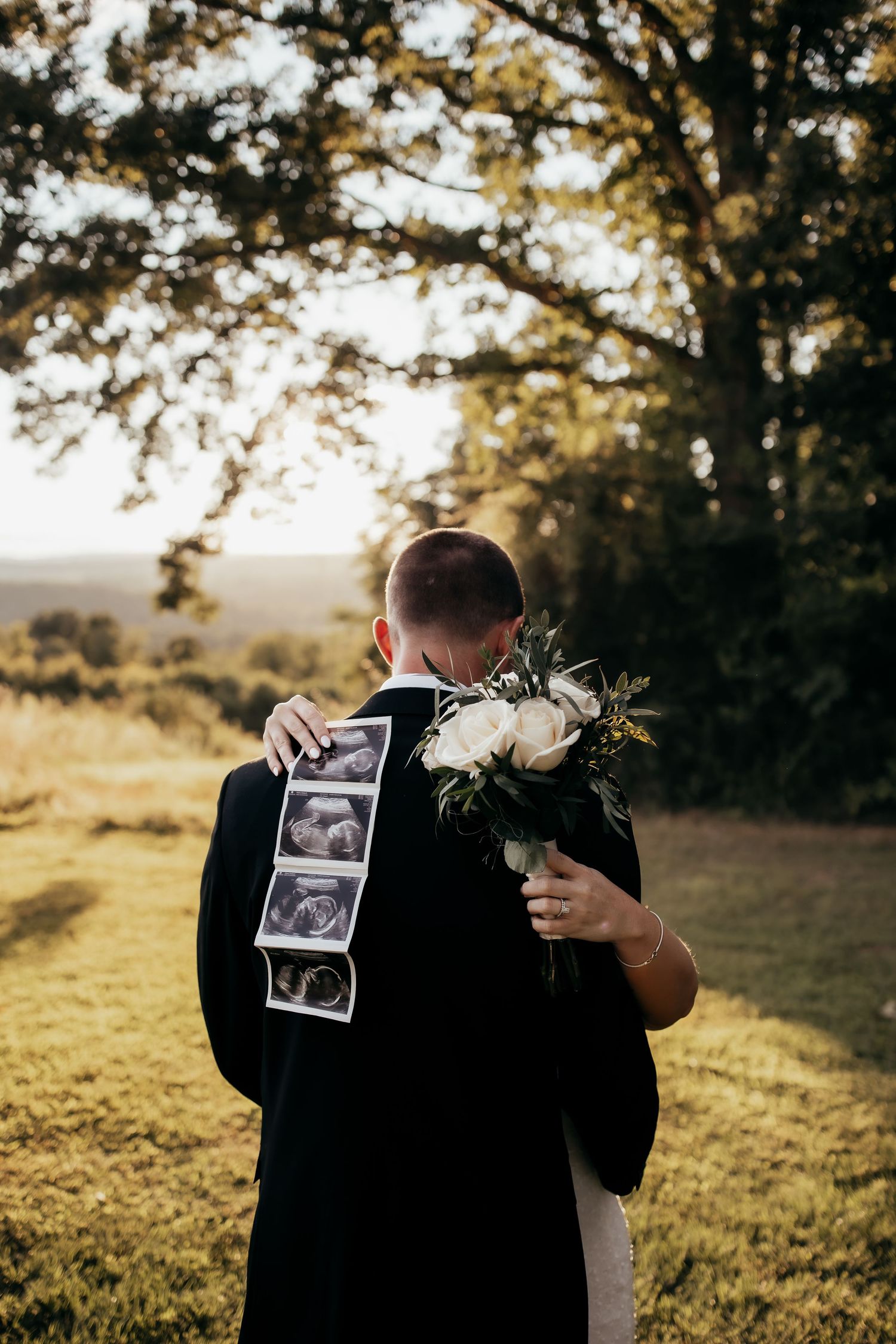 Bride and groom embracing outdoors, holding ultrasound photos and white floral bouquet, announcing pregnancy on wedding day.