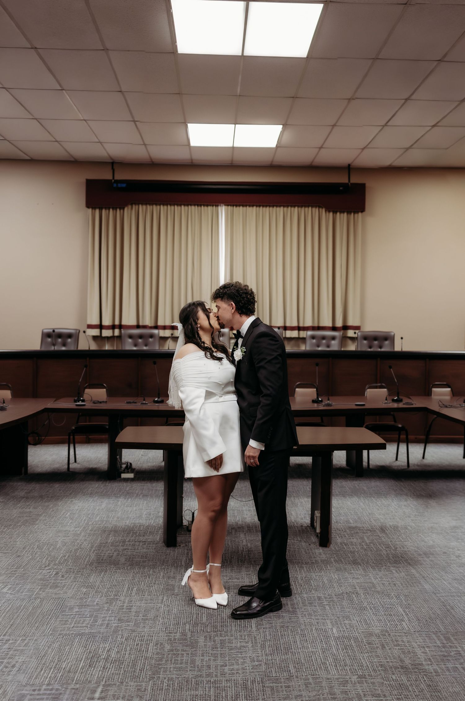 Bride and groom kissing in empty courthouse boardroom, bride in white mini dress holding a bouquet.