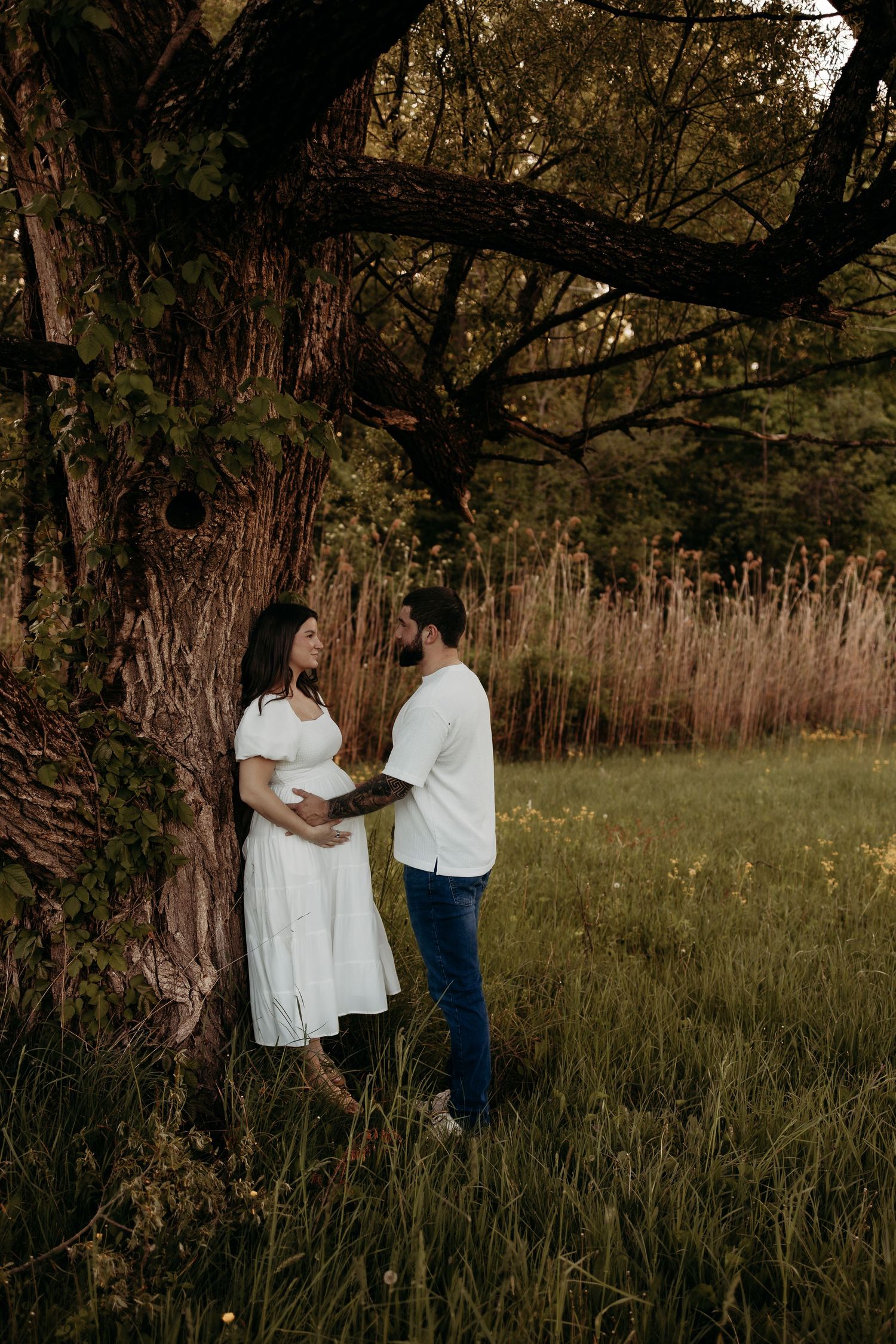 Pregnant couple sharing a tender moment under a large tree in a lush green field, woman in white dress cradling her baby bump.