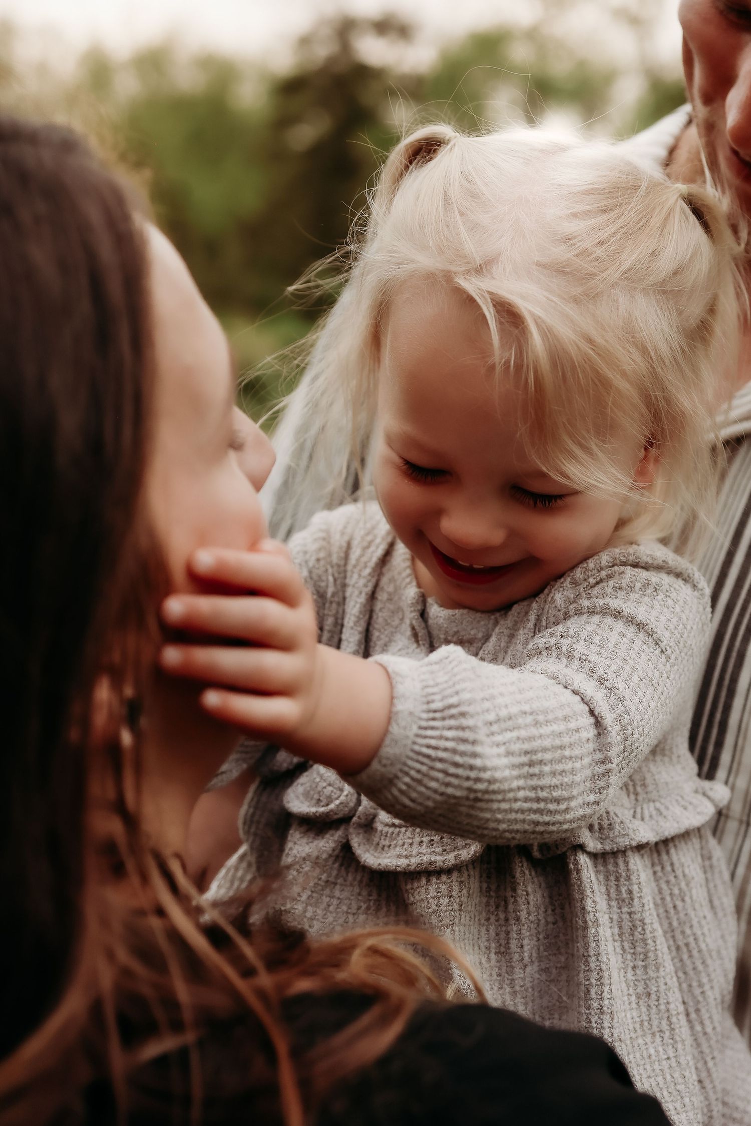 Laughing toddler girl with blonde hair reaches toward her mother in a sweet outdoor family moment.
