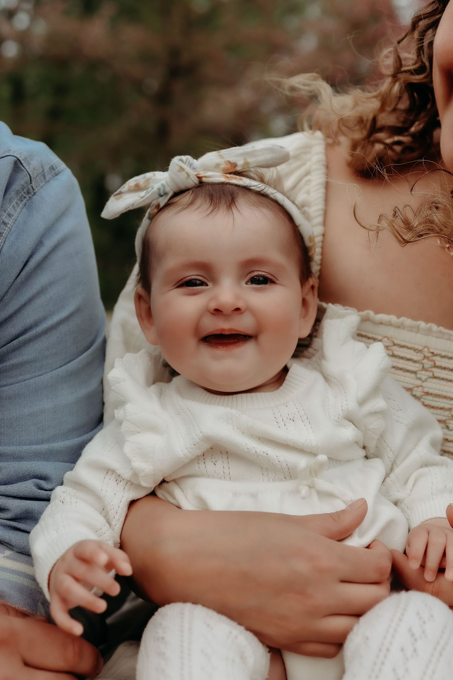 Adorable smiling baby in white knit outfit with floral headband sitting with parents outdoors.