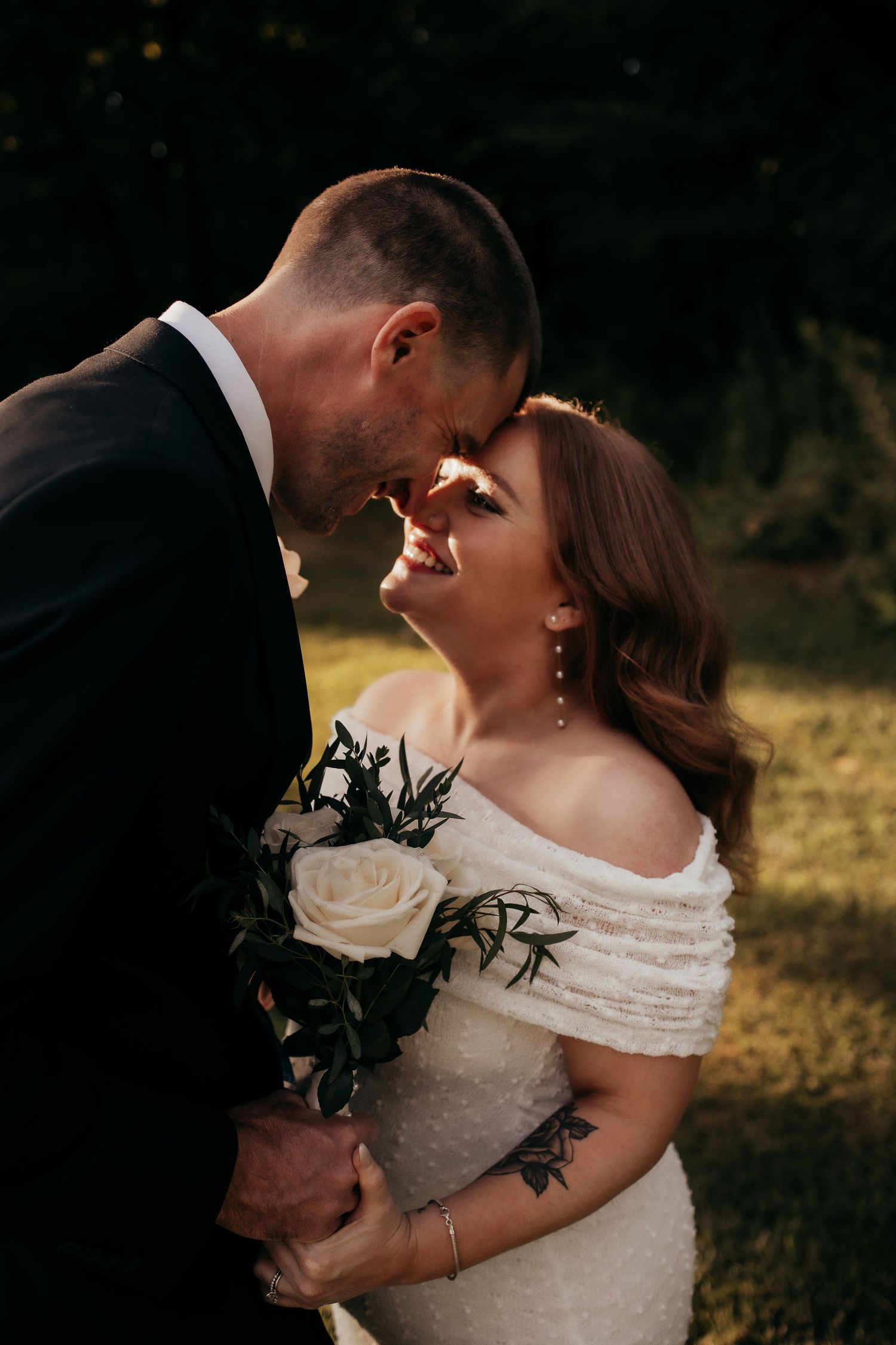 A bride and groom share an intimate moment, foreheads touching, as she holds a white rose bouquet in golden sunlight.