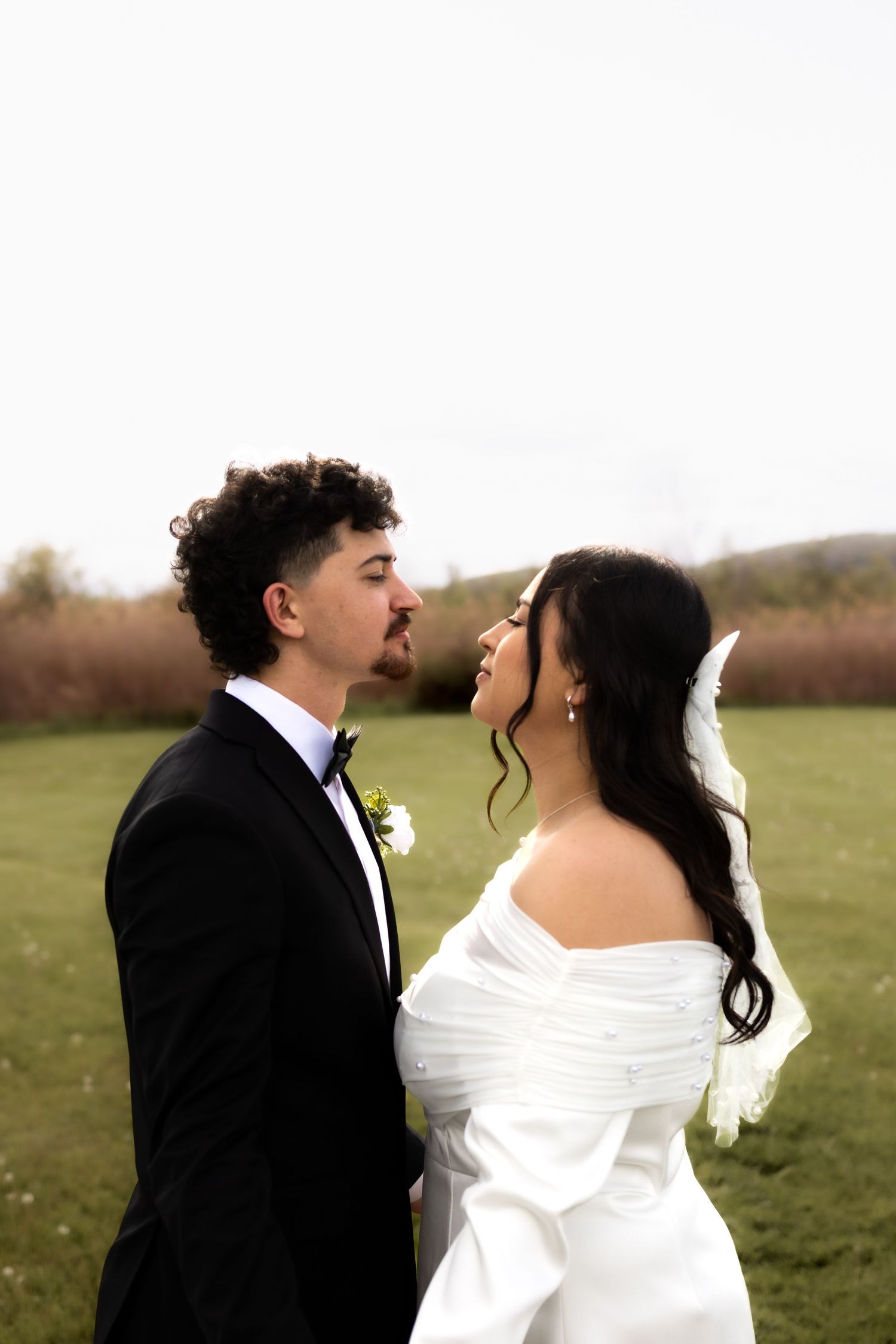 A bride and groom share a romantic moment outdoors, dressed in a white off-shoulder gown and black tuxedo.