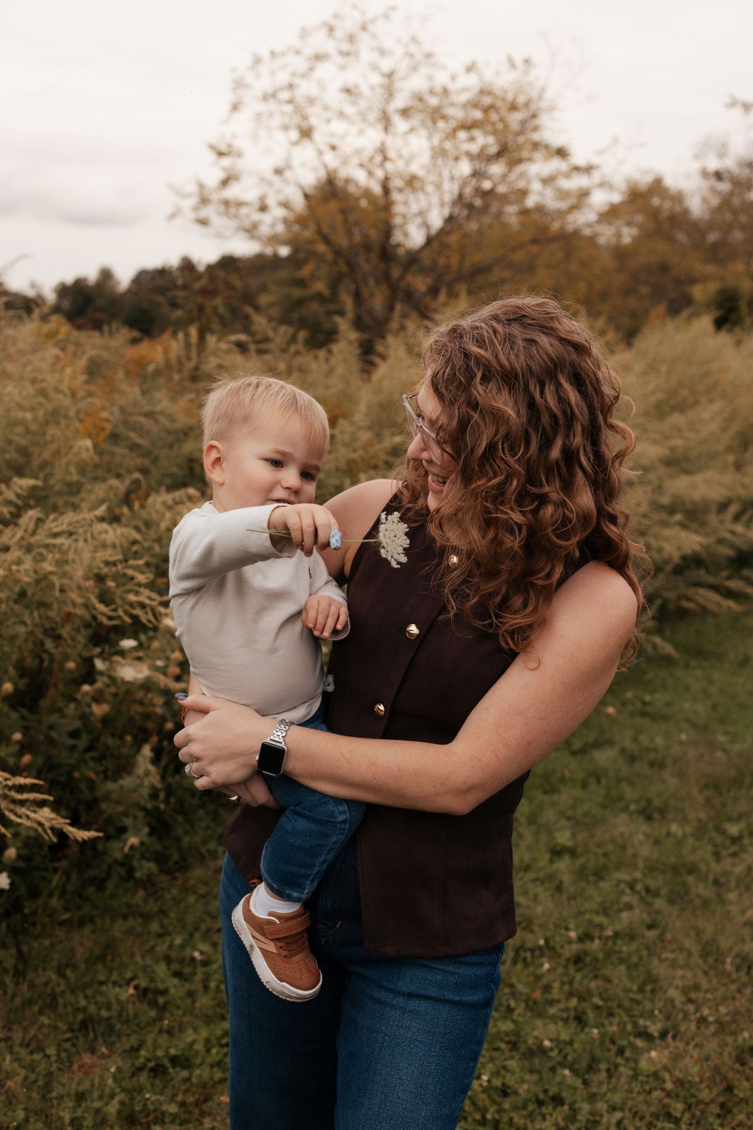 A curly-haired woman in a brown vest holds a smiling baby boy outdoors in a lush green autumn setting.