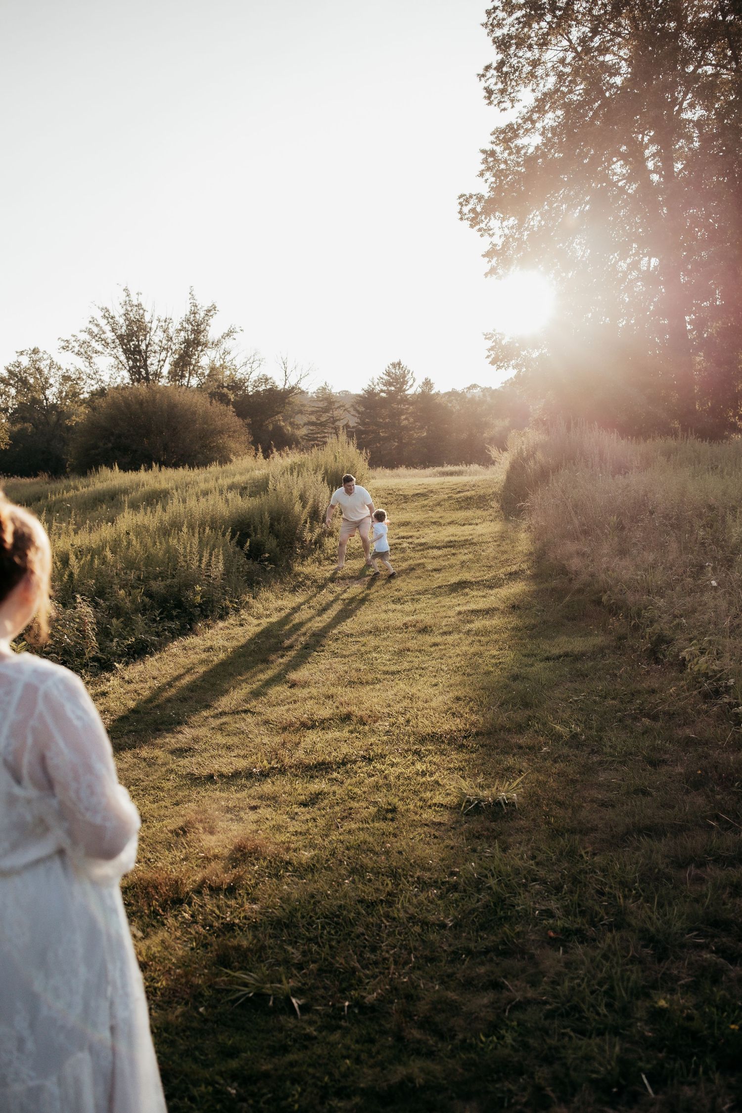 Family running through a sunlit meadow at golden hour, surrounded by lush greenery and tall trees.