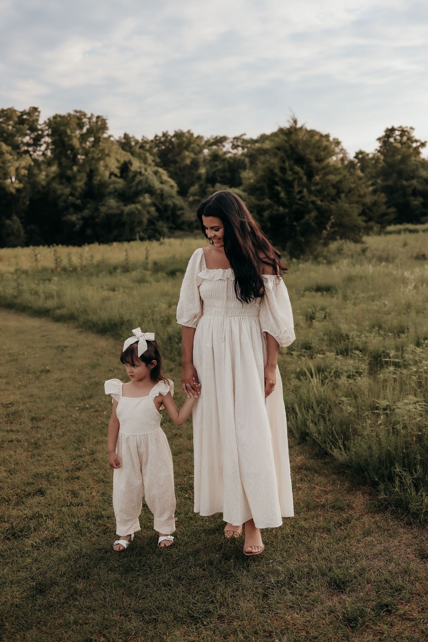 Mother and young daughter in matching white outfits walking in green meadow at dusk.