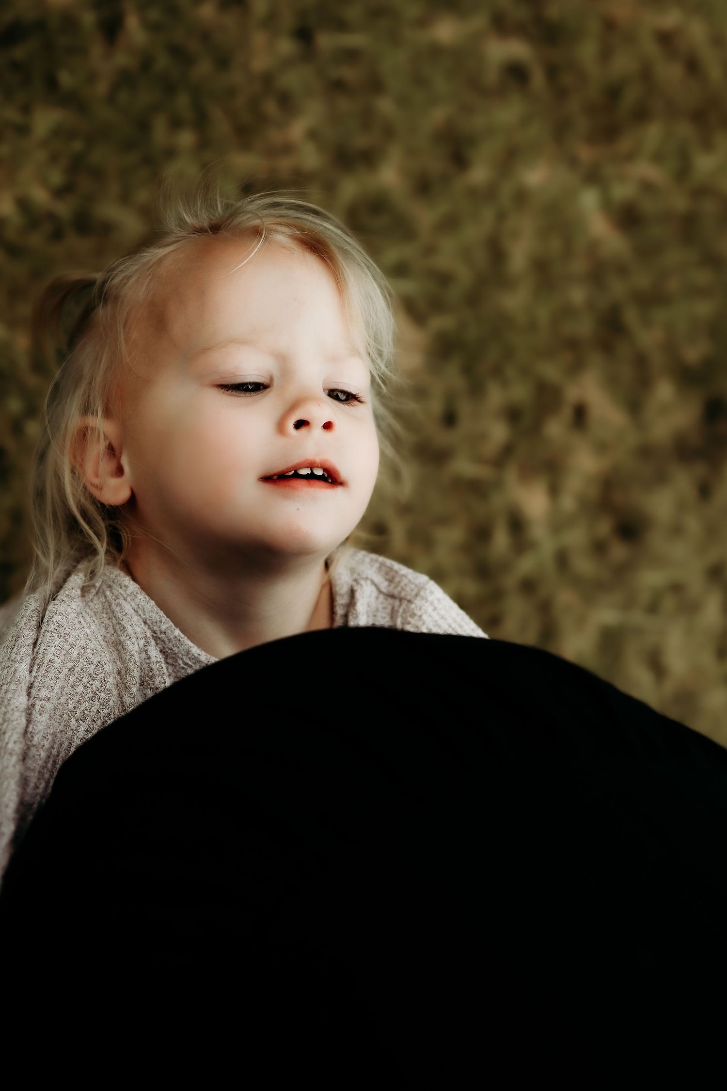 Adorable blonde toddler girl with a sweet smile peeking over a dark object in a natural outdoor setting.