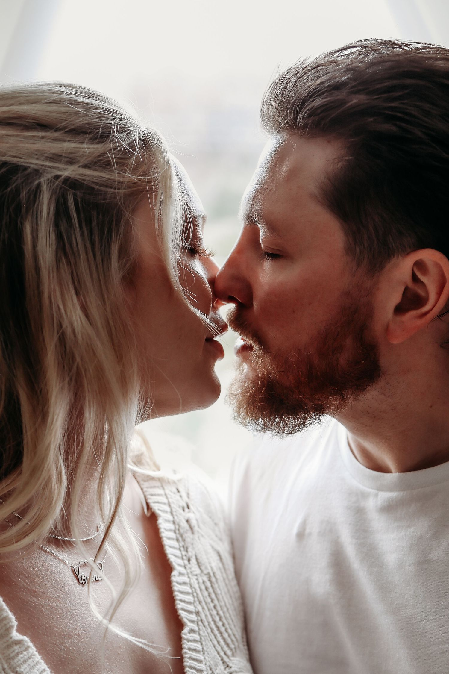 Romantic close-up of a bearded man and blonde woman sharing a kiss near a bright window.