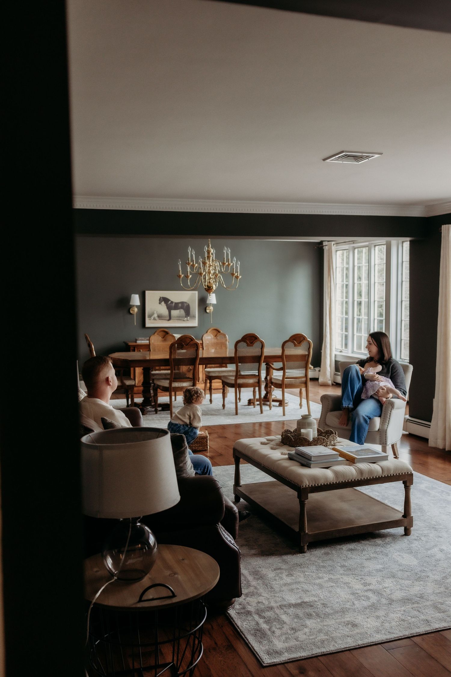 Family relaxing in an elegant dark-walled living room with chandelier, hardwood floors, and large windows.