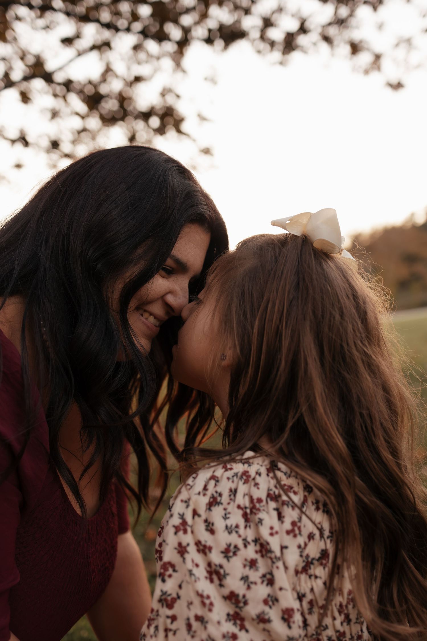 A smiling mother with long dark hair touches noses with her daughter, who wears a white bow and floral top, outdoors at golden hour.