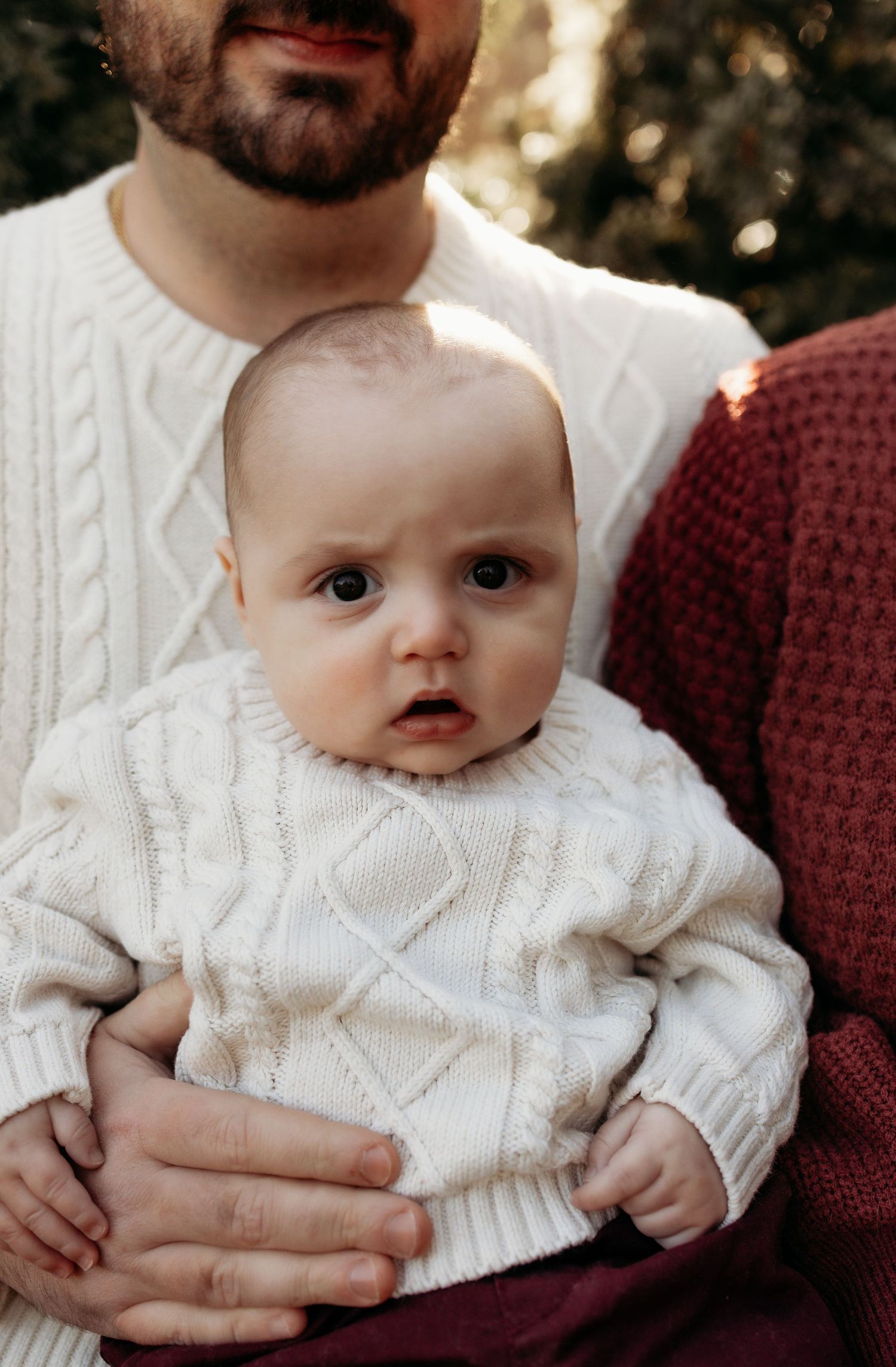 A baby in a white cable-knit sweater is held by two adults in cozy sweaters outdoors.