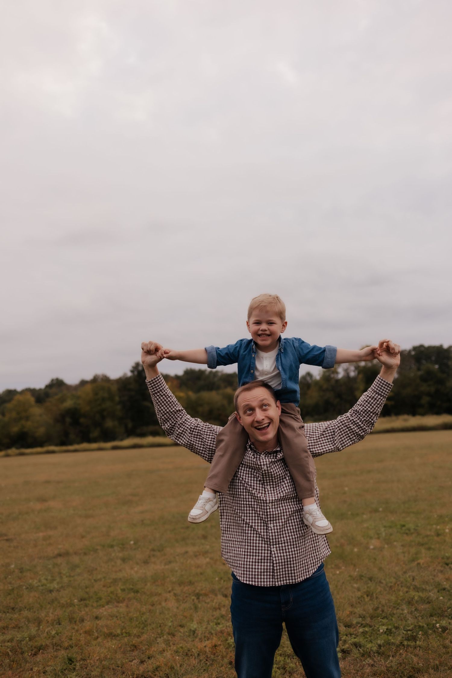A joyful father carries his laughing young son on his shoulders in an open green field on a cloudy day.
