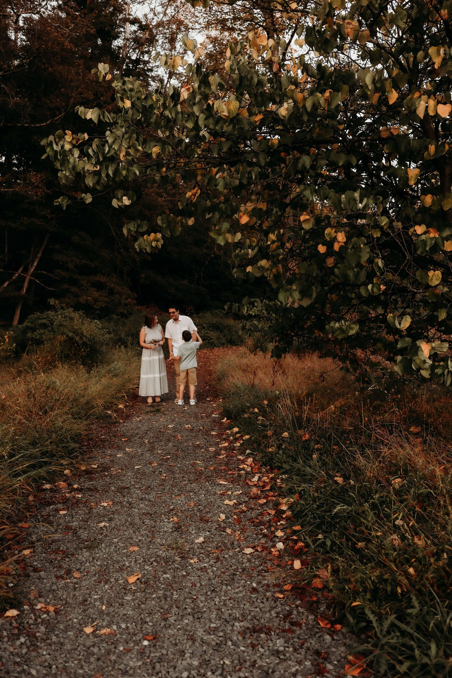 Family of three walking on a gravel path surrounded by autumn foliage and trees in a serene forest setting.
