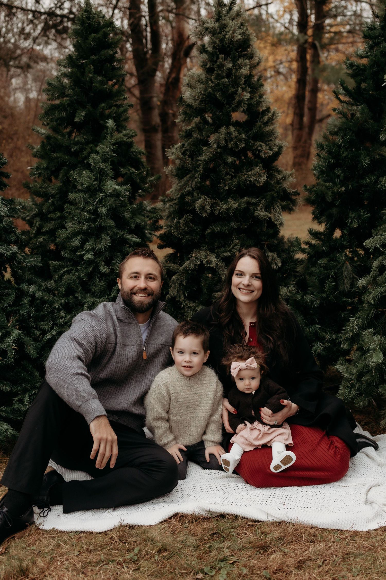 Family of four posing on white blanket surrounded by evergreen trees for a festive holiday photo session.